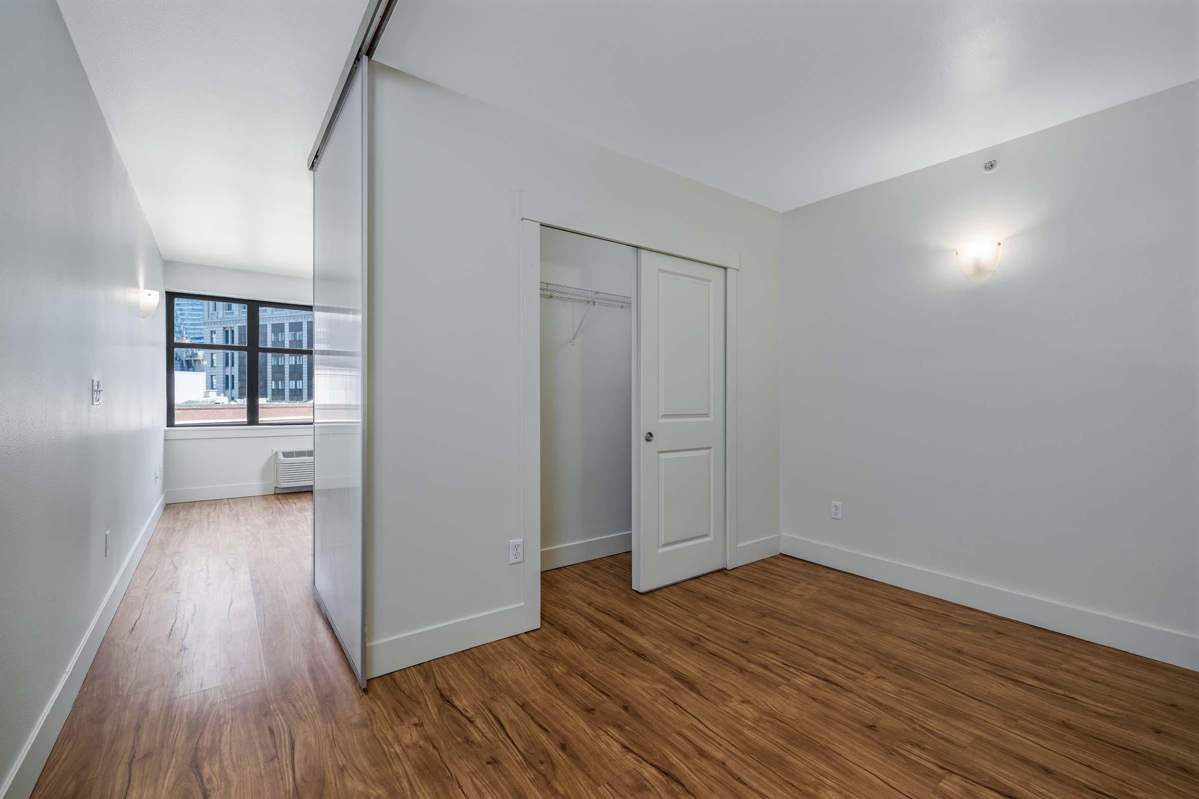 Interior view of a modern, empty room with light wooden flooring, white walls, and a large window. There is a small closet with sliding doors, and a stylish light fixture on the wall. The room has a clean and minimalistic design, suitable for various furniture arrangements.