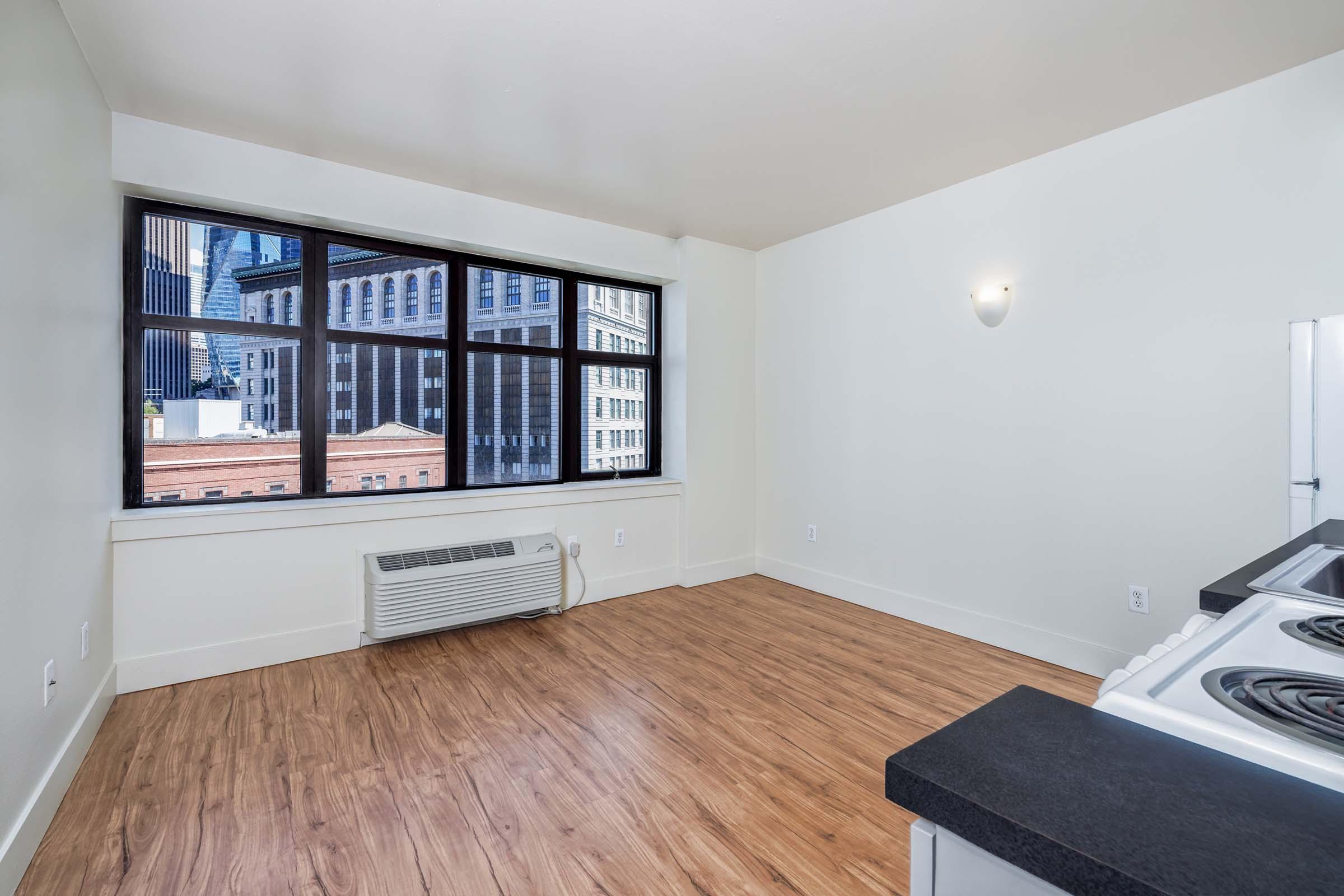 A brightly lit room with large windows showing a cityscape, featuring a wooden floor and a kitchenette with a stove. The walls are painted white, and there is an air conditioning unit on the wall.