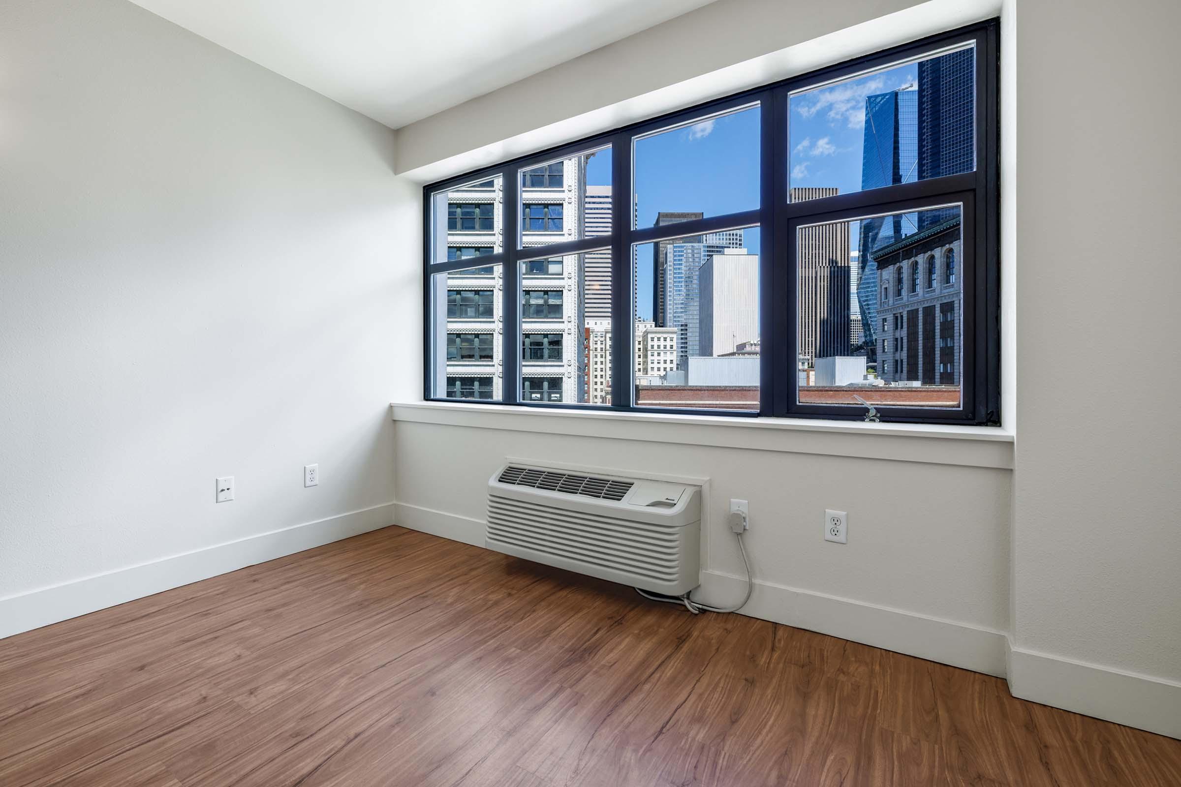 Empty room with large windows showcasing a city skyline. The walls are painted light colors, and the flooring is wood-like laminate. An air conditioning unit is installed under the window, emphasizing the modern and minimalistic design of the space.
