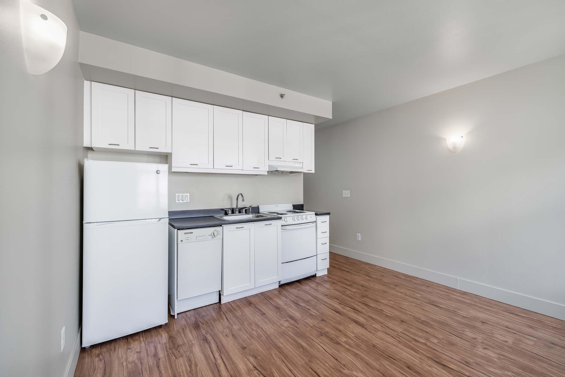 A modern kitchen with white cabinets and appliances, including a refrigerator, dishwasher, and stove. The flooring is light wood, and the walls are painted in a neutral color. Natural light comes in, and there is a wall-mounted light fixture. The space is clean and uncluttered, ideal for a small apartment.