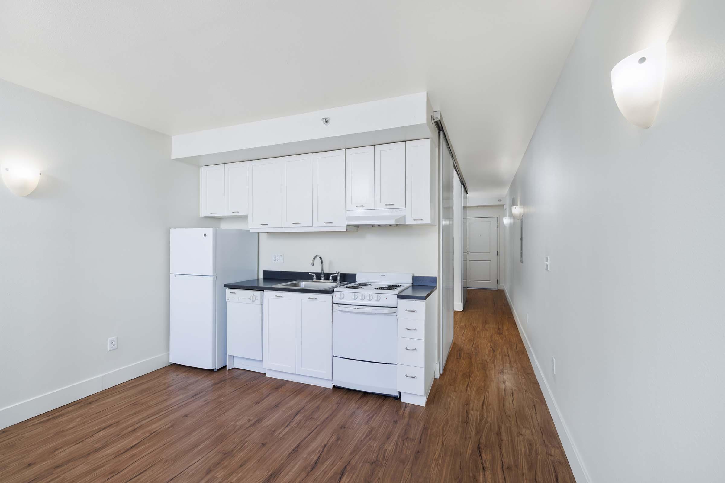 A bright, unfurnished studio apartment featuring a modern kitchen area with white cabinets, a refrigerator, dishwasher, and stove. The space has light-colored walls and hardwood flooring, with soft lighting fixtures on the walls. A hallway leads to an open door in the background.