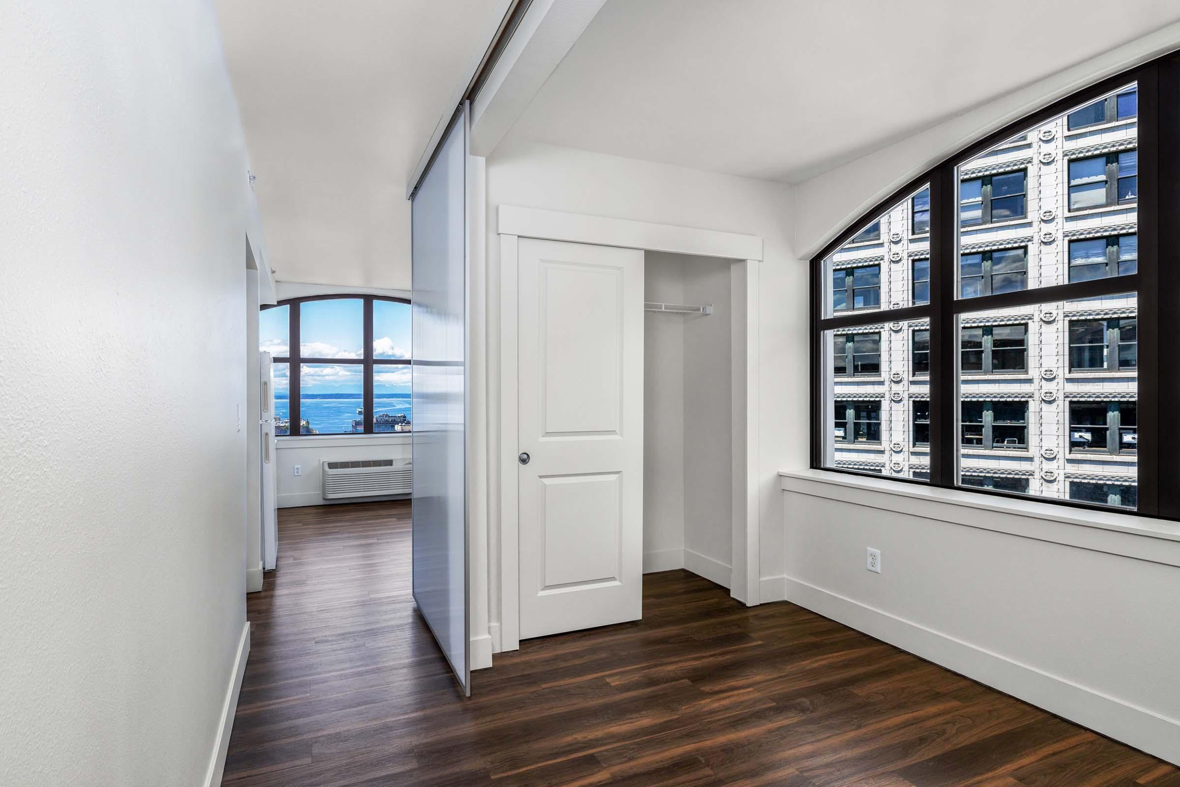 A bright, modern apartment interior featuring a spacious entryway with white walls, hardwood flooring, and large windows showcasing a view of a cityscape. A sliding door leads to a closet area, while an air conditioning unit is visible along one wall. Natural light floods the space, creating an inviting atmosphere.