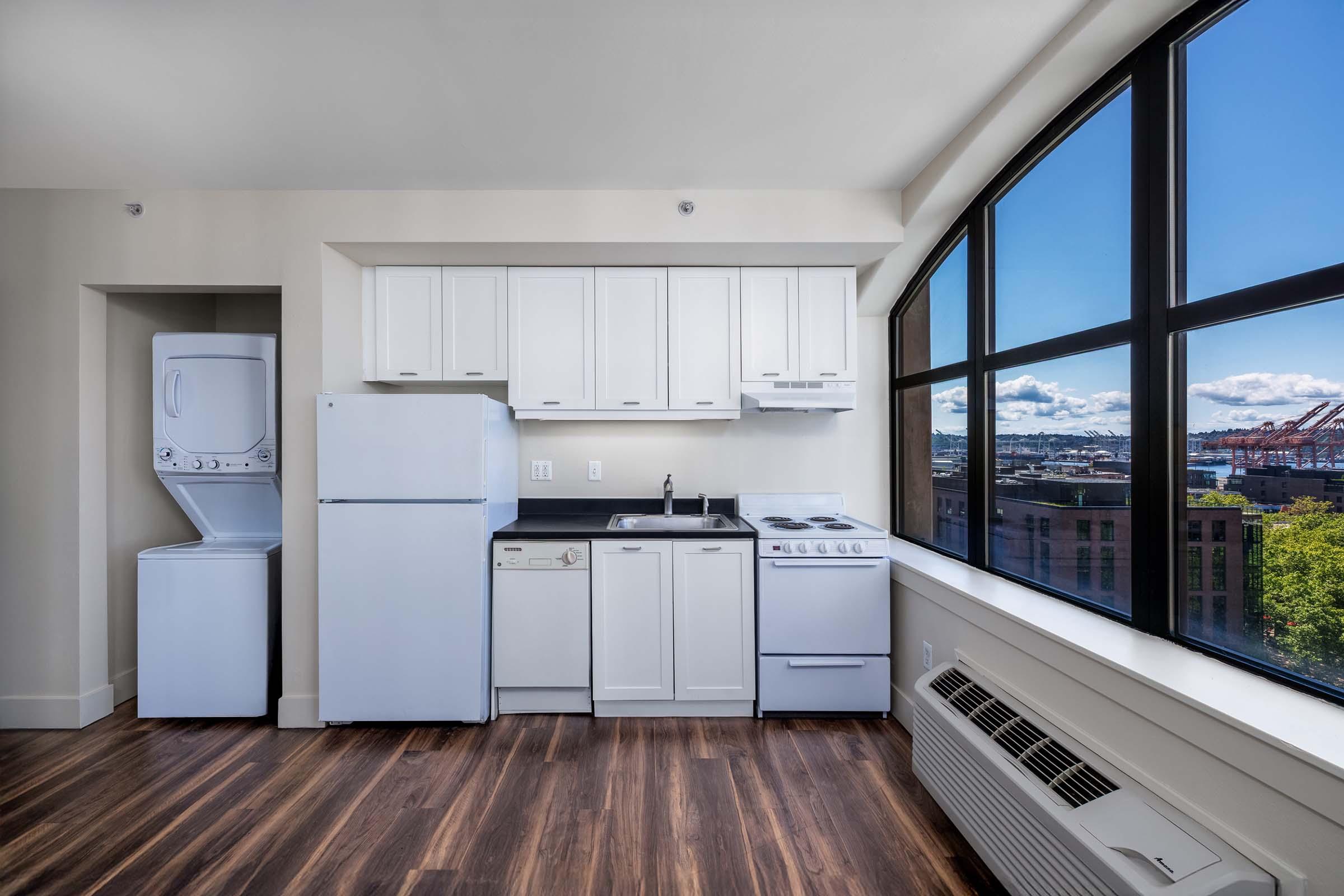Bright kitchen area with modern white cabinetry, a refrigerator, dishwasher, and gas stove. A washer and dryer unit is located next to the kitchen. Floor-to-ceiling windows provide ample natural light and a view of the city skyline and clouds. The flooring is dark wood, adding a warm contrast to the space.