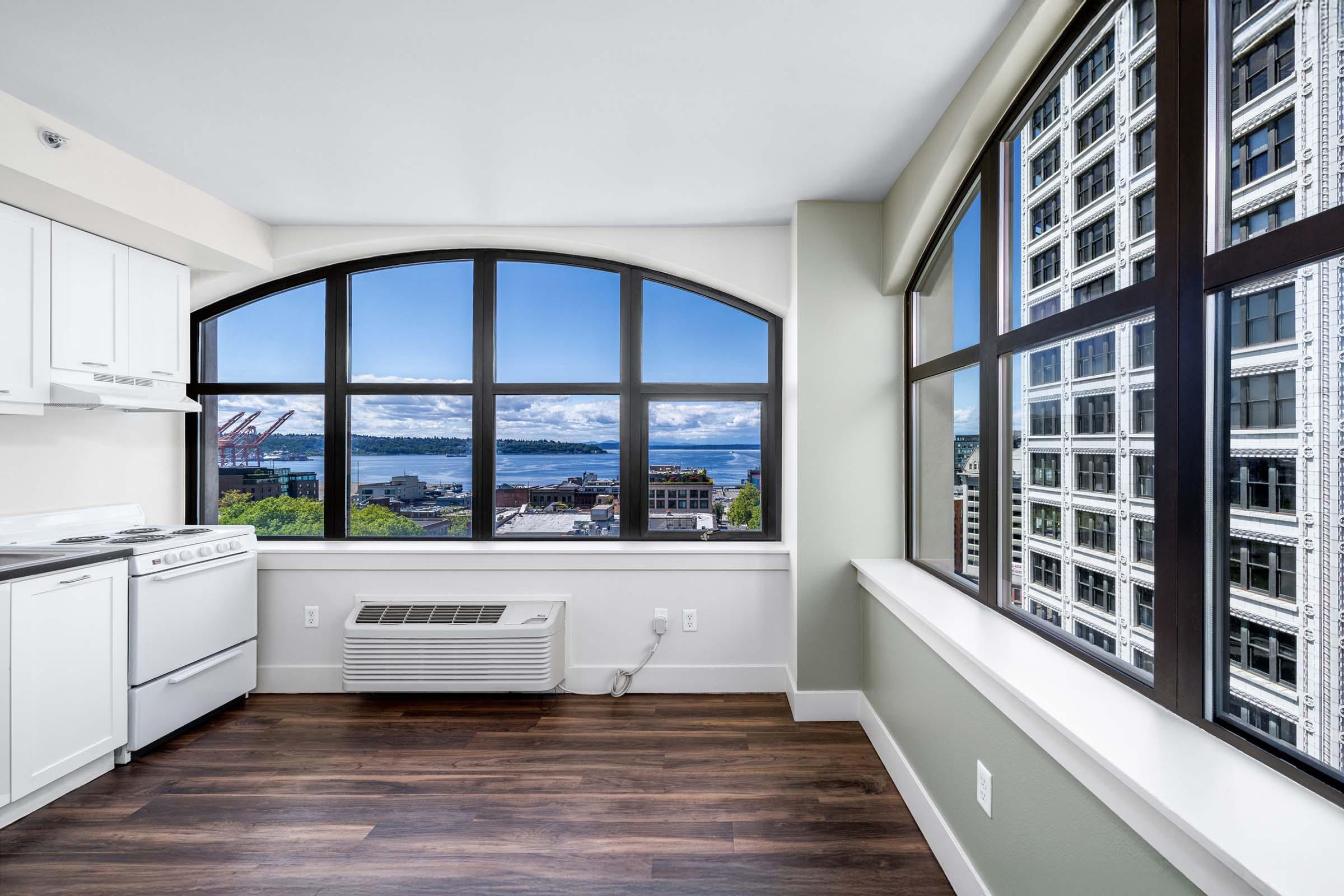 Interior view of a modern kitchen and living space featuring large windows that showcase a scenic view of the skyline and harbor. Light walls and wooden flooring create a bright, airy atmosphere. The room is well-lit with natural light, highlighting a clean, contemporary design.