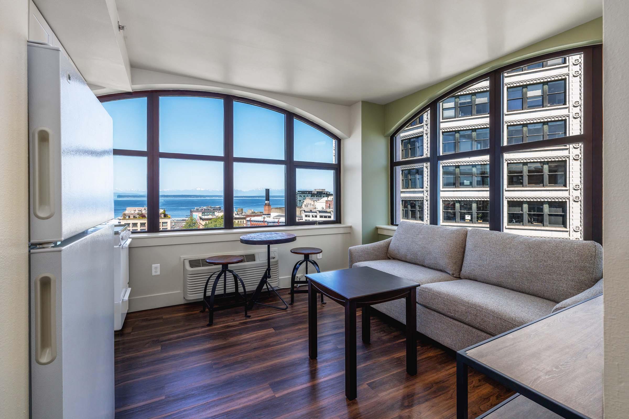A cozy living space featuring a light-colored couch, a small wooden table, and a refrigerator. Large arched windows provide a scenic view of the ocean and city buildings, allowing natural light to fill the room. The flooring is dark wood, creating a warm ambiance.