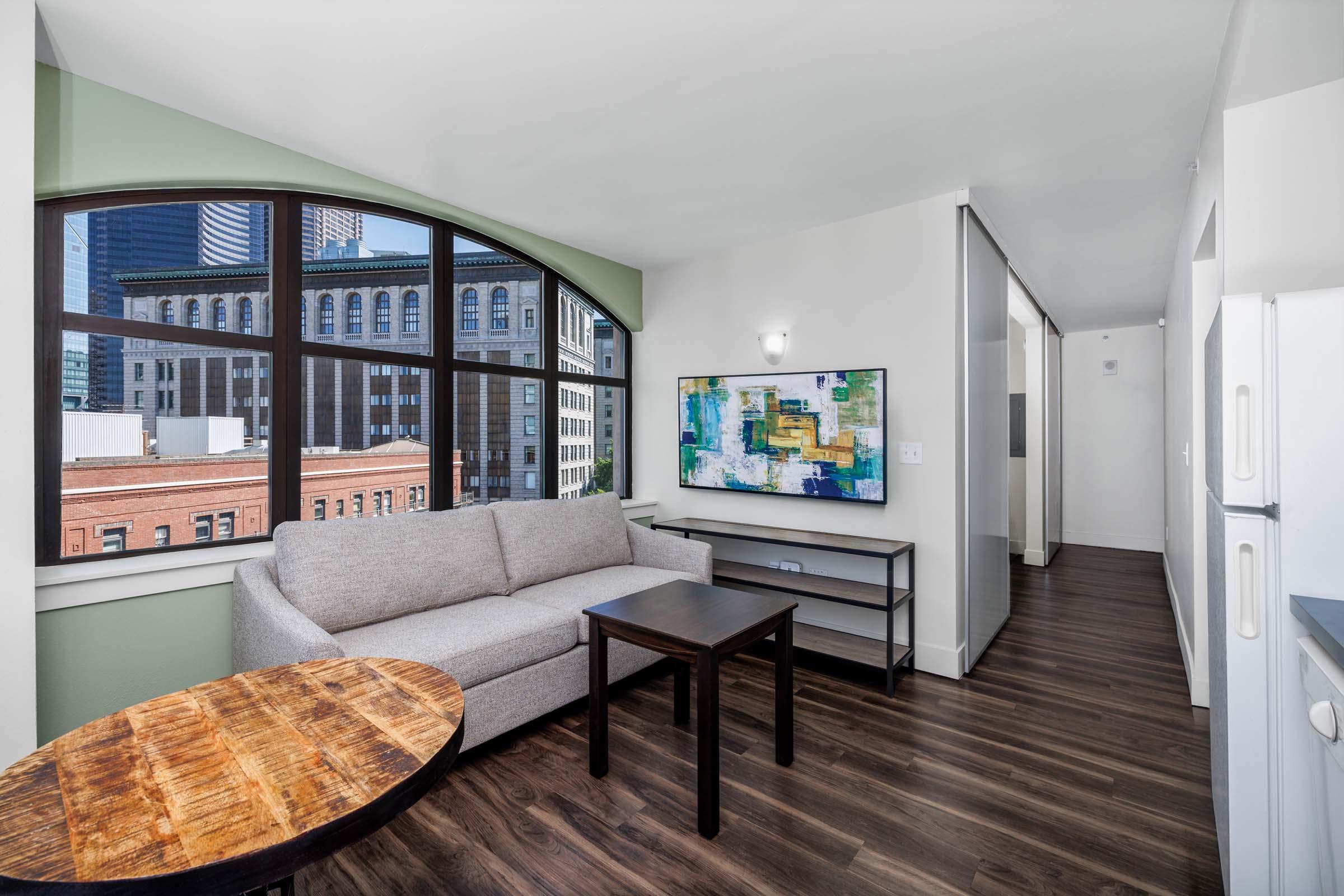 A modern, well-lit living area featuring a gray sofa and a wooden coffee table. A colorful abstract painting hangs on the wall, and large windows provide a view of the cityscape. The room has a contemporary design with dark wood flooring and a hallway leading to other areas.