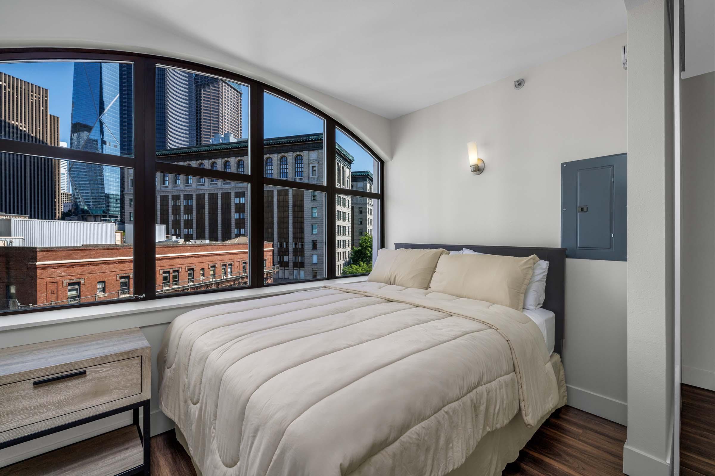 A cozy bedroom with a large window offering a view of city skyscrapers. The room features a neatly made bed with beige bedding, a bedside table, and a clean, modern aesthetic. Sunlight brightens the space, creating a welcoming atmosphere.