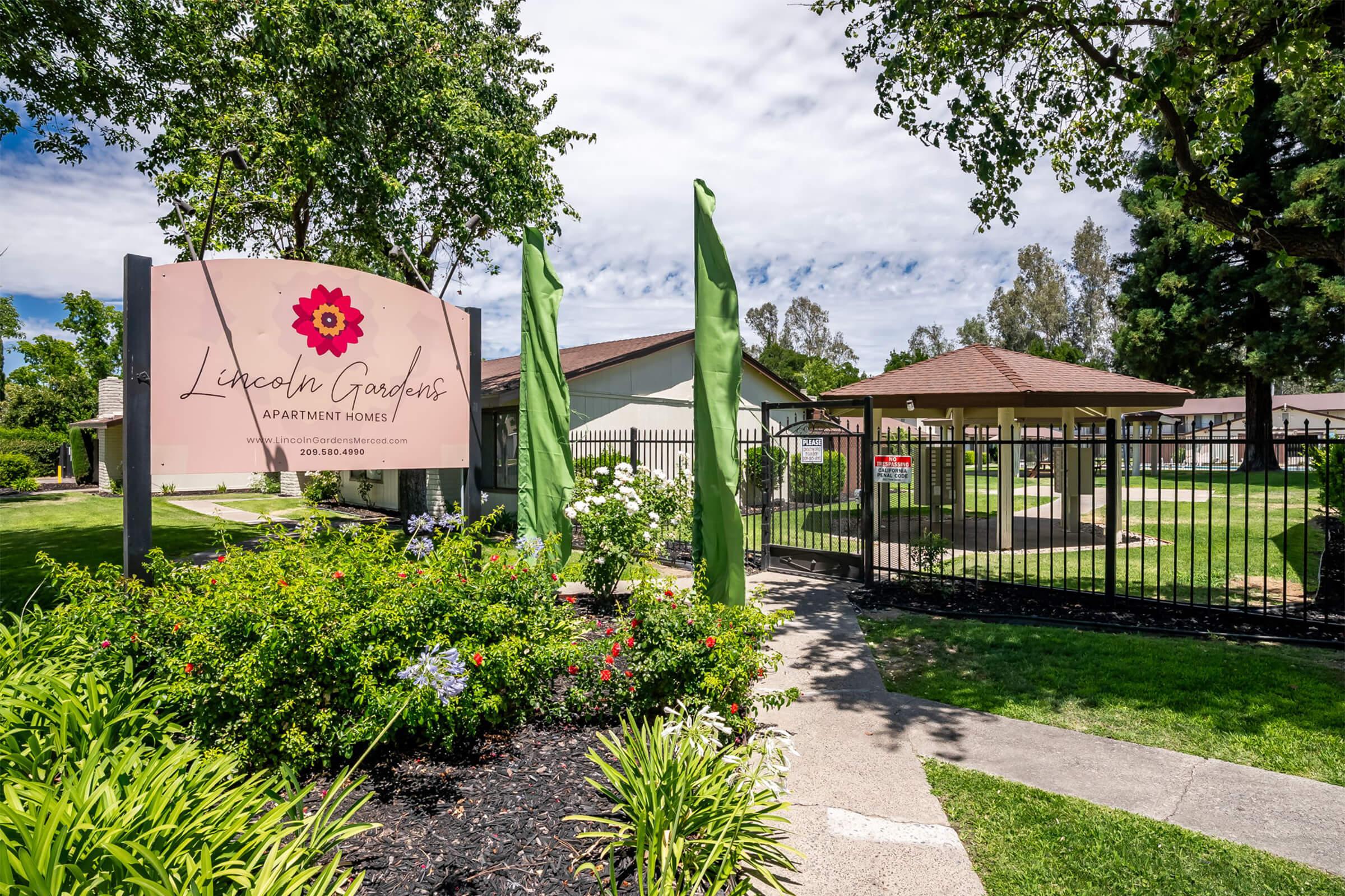 Sign for Lincoln Gardens Apartment Homes with decorative flowers and greenery in the foreground, featuring a fenced area with a gazebo and well-maintained landscaping. The sky is partly cloudy, and the surroundings include trees and manicured lawns.