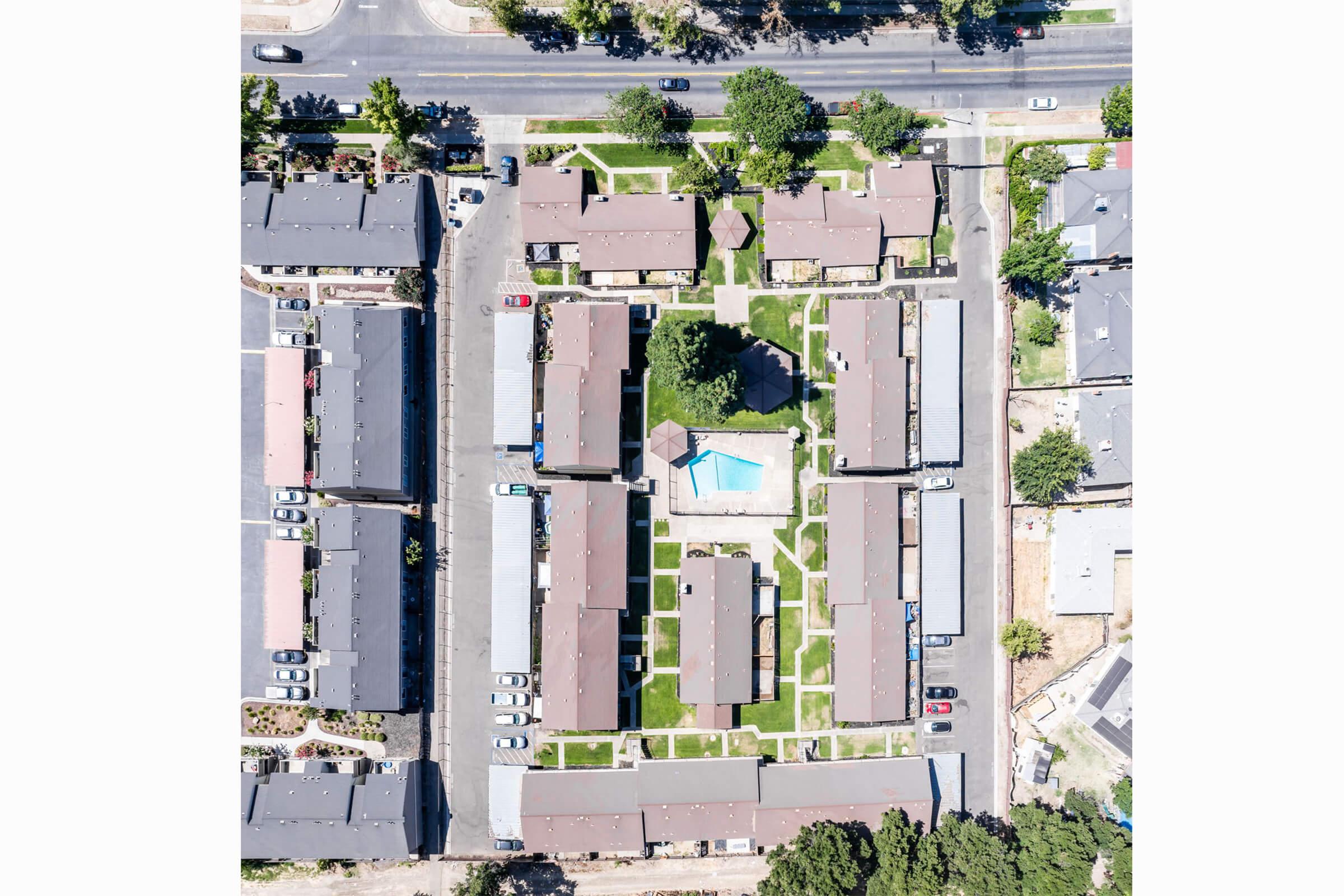 Aerial view of a residential complex featuring multiple single-story buildings arranged in a rectangular layout around a central grassy area with a small pool. Surrounding the complex are streets lined with vehicles and trees, indicating a suburban environment.