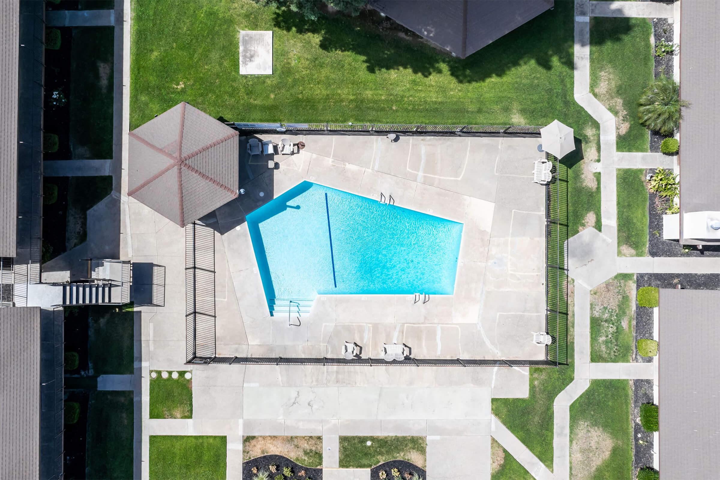Aerial view of a swimming pool surrounded by concrete decking and green grass. The pool features a unique shape with steps leading in, and there are lounge chairs arranged around it. Two shaded pavilions are visible near the pool, along with paved walkways and landscaped areas.
