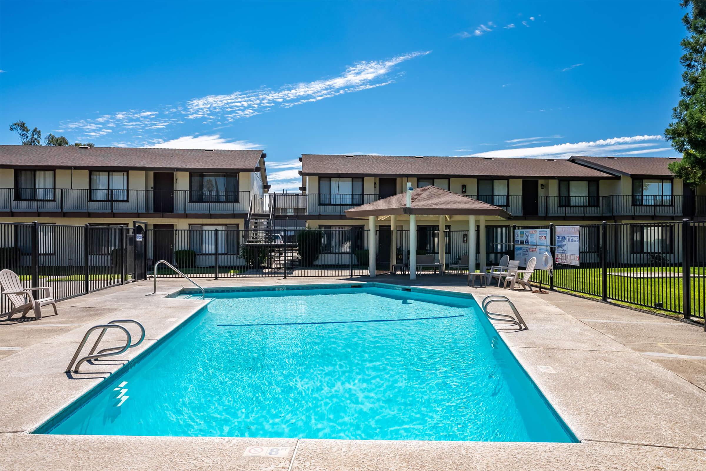A clear blue swimming pool surrounded by a concrete deck, with lounge chairs and a gazebo nearby. In the background, two-story apartment buildings with balconies are visible, along with a green lawn and a blue sky with wispy clouds. The area is fenced for privacy.