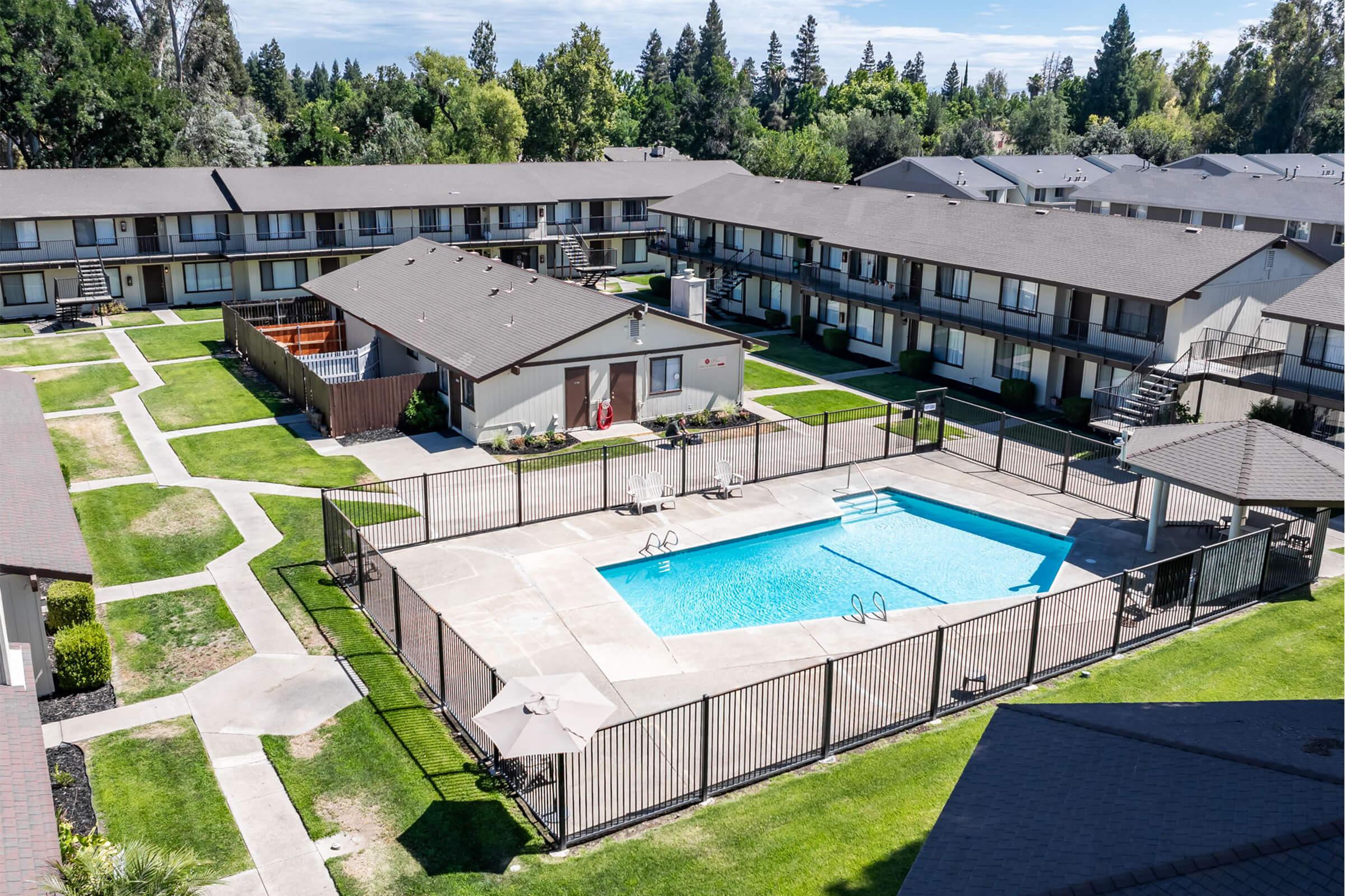 Aerial view of a residential complex featuring multiple single-story buildings arranged around a central swimming pool. The pool is enclosed by a fence, with lounge chairs and umbrellas nearby. Green lawns and pathways connect the buildings, surrounded by trees in the background under a clear blue sky.
