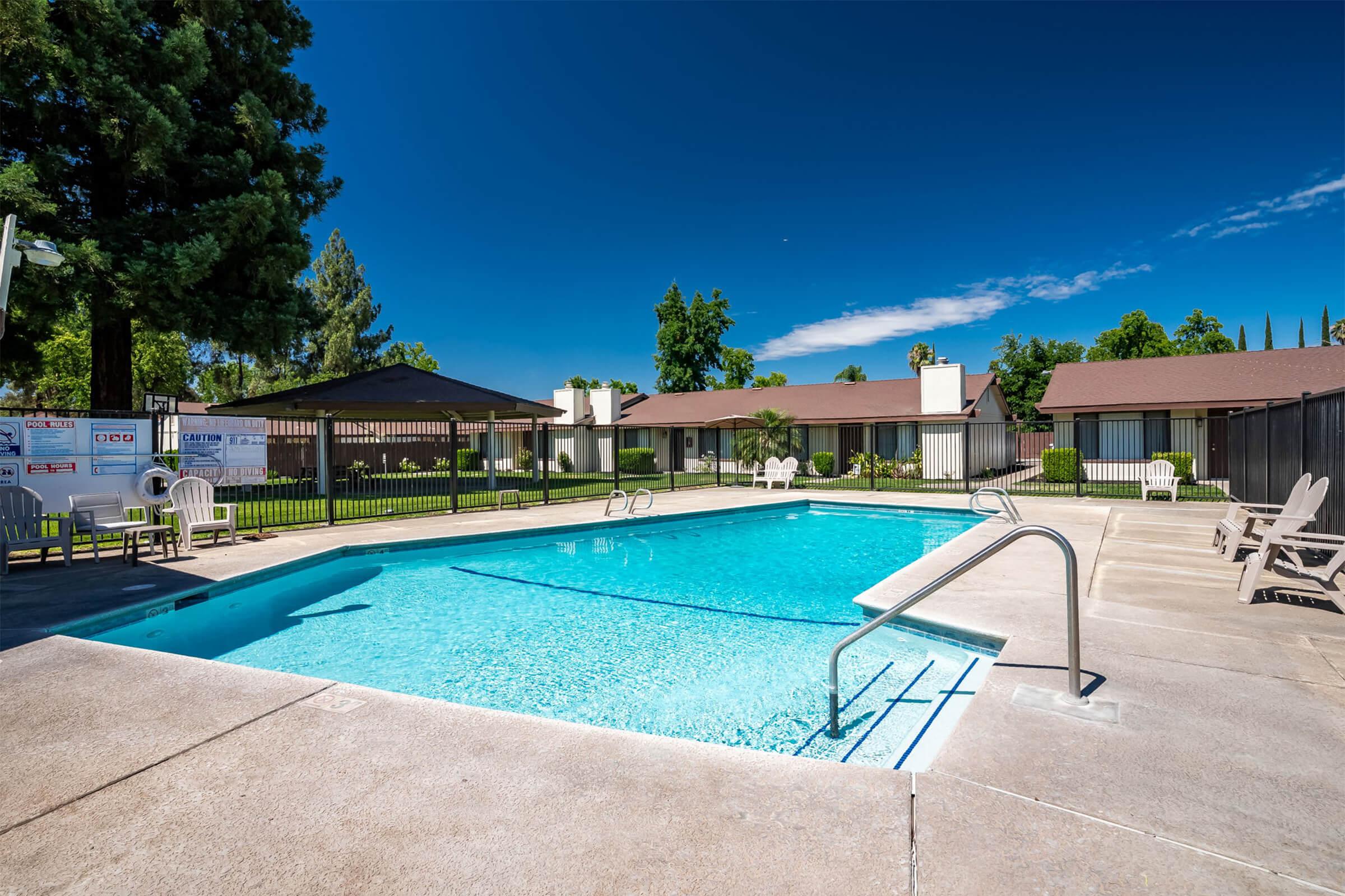 A clear blue swimming pool surrounded by lounge chairs, with a backdrop of green trees and a bright blue sky. There are white picket fences and a few buildings in the distance, creating a relaxing outdoor atmosphere.