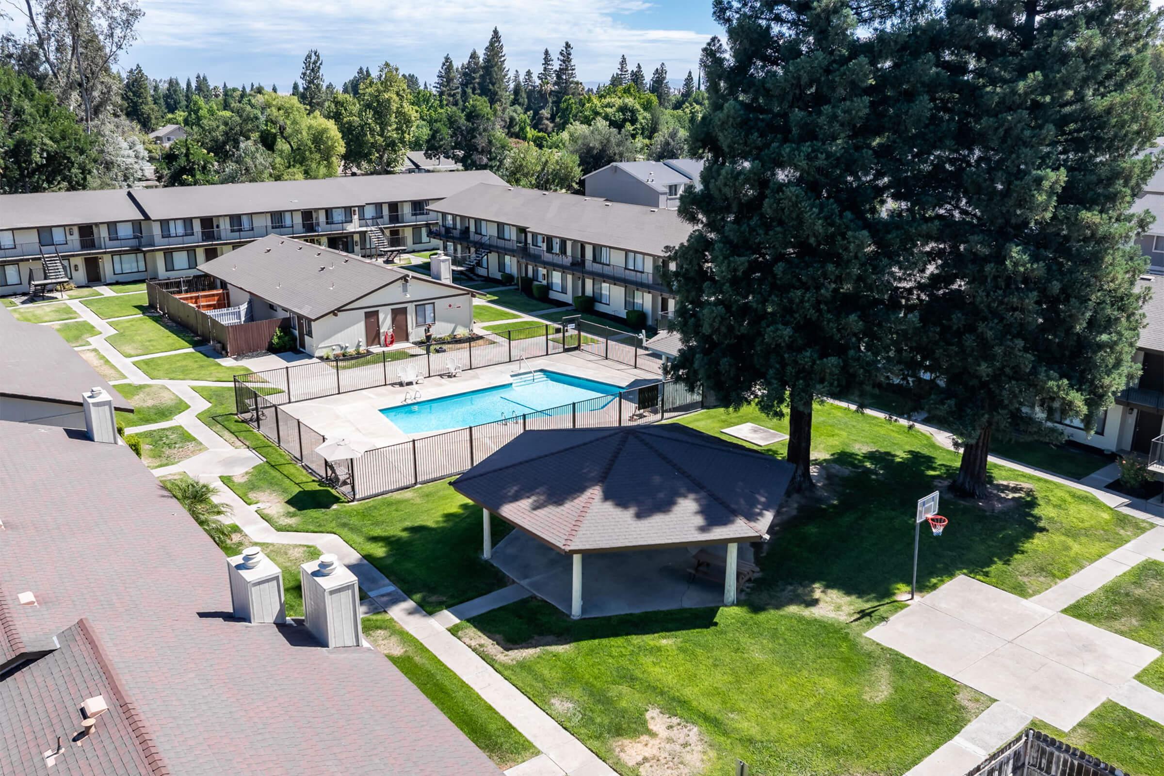 Aerial view of a residential complex with multiple buildings surrounding a swimming pool. The area features landscaped lawns, a gazebo, and a basketball hoop, set against a backdrop of tall trees and a clear blue sky. The scene conveys a peaceful and inviting atmosphere for residents.