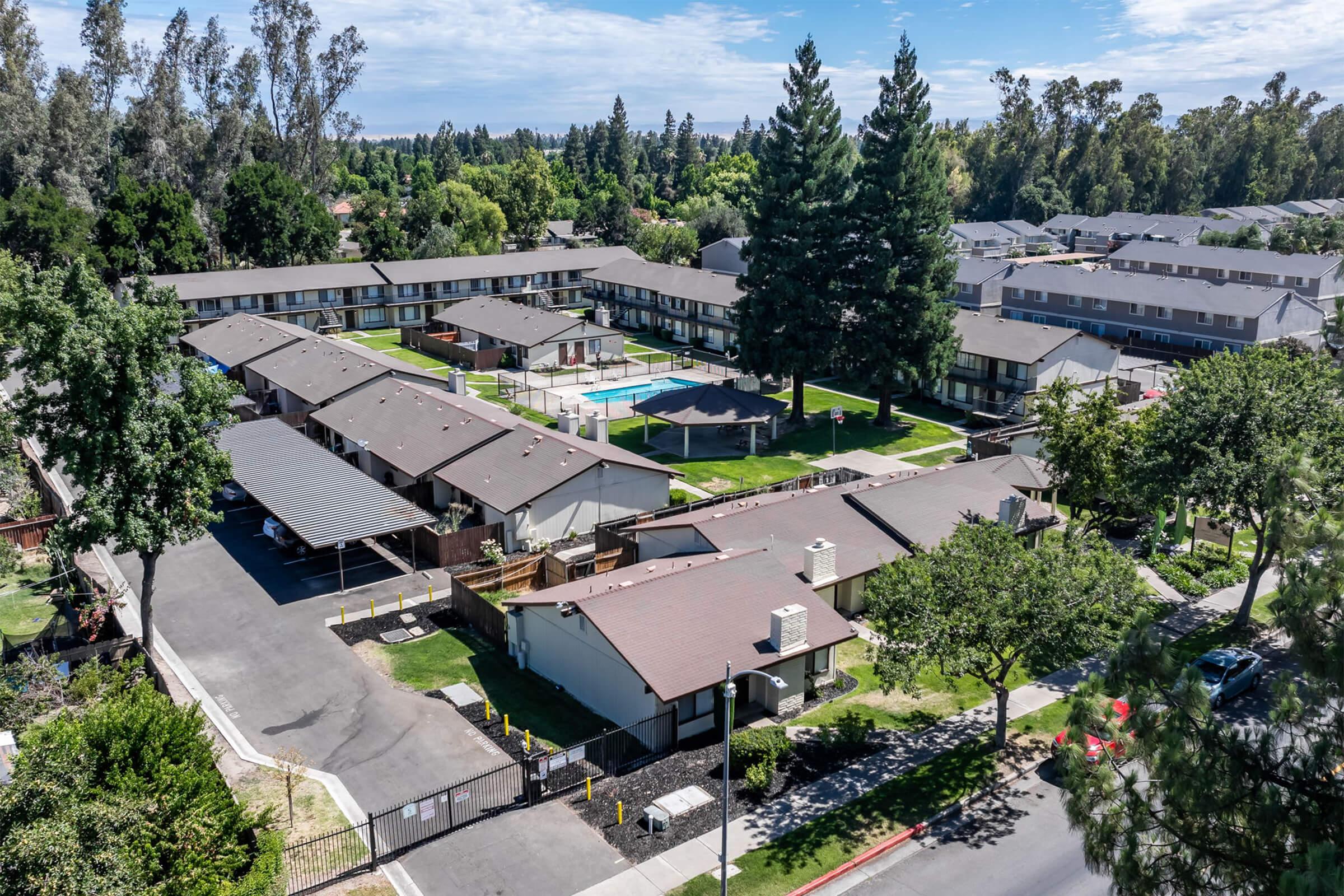 Aerial view of a residential apartment complex featuring multiple buildings arranged around a central courtyard. The area includes landscaping with trees, a swimming pool, and parking spaces. Blue sky and scattered clouds are visible in the background, indicating a sunny day.