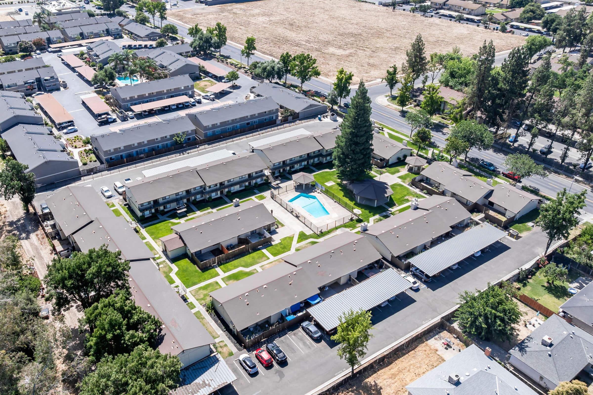 An aerial view of a residential complex featuring several small apartment buildings arranged around a central pool and green space. Surrounding the complex are trees and parking areas, with streets visible in the background. The setting appears well-maintained and organized.