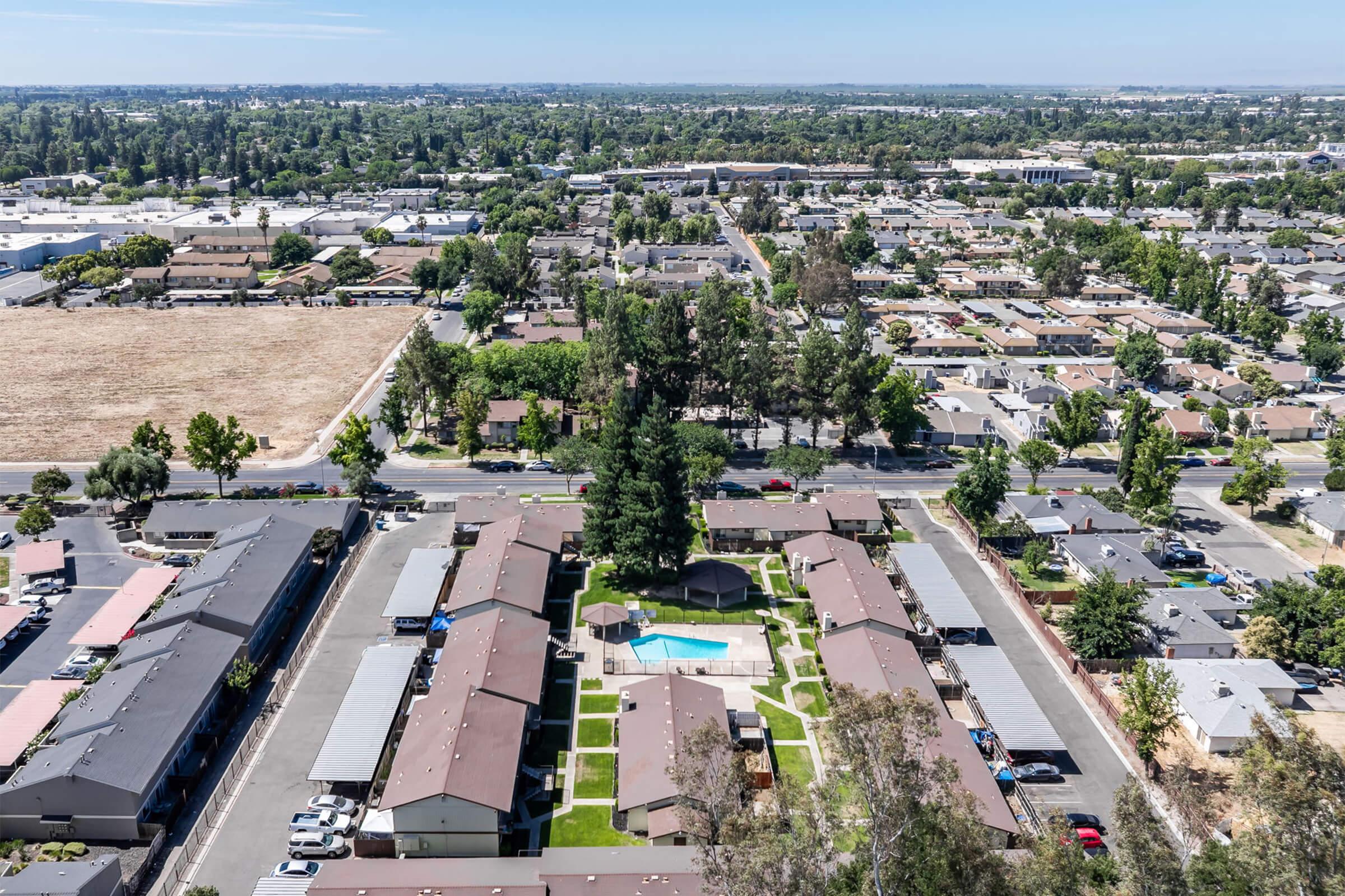 Aerial view of a residential area featuring several buildings, with a central courtyard and swimming pool. Surrounding the complex are trees, roads, and additional homes. In the background, there are open fields and a mix of urban development, showcasing a blend of greenery and residential structures.