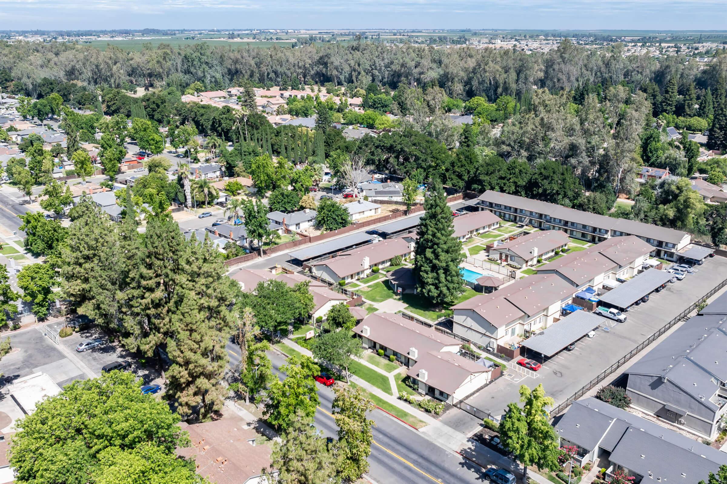 Aerial view of a suburban neighborhood featuring residential buildings, tree-lined streets, and green spaces. The area includes multi-family homes, individual houses, and well-maintained lawns, with a mix of trees providing shade. The landscape appears lush and developed, reflecting a typical family-friendly community.