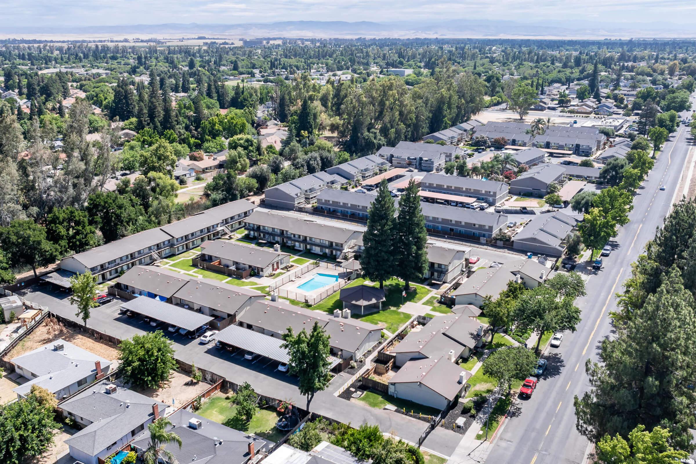 Aerial view of a residential area featuring multiple apartment buildings arranged around a central green space with a swimming pool. Surrounding trees and streets are visible, along with a mix of single-story and multi-story structures. The scene is set under a clear blue sky.