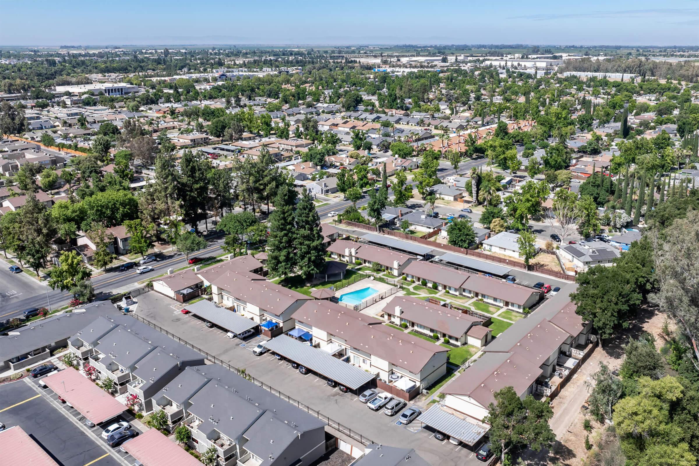 Aerial view of a residential neighborhood featuring multiple low-rise buildings surrounding a central swimming pool. The area is lush with trees and greenery, and nearby streets are visible, lined with houses and parked cars. In the background, industrial buildings and open land can be seen.