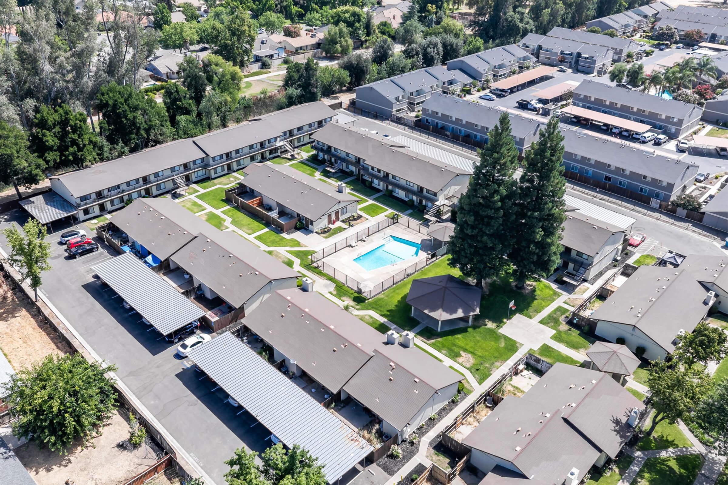Aerial view of a residential complex featuring several single-story buildings arranged around a central courtyard with a swimming pool. The area is landscaped with green lawns, trees, and shaded parking spaces. Surrounding residential buildings and greenery can be seen in the background.
