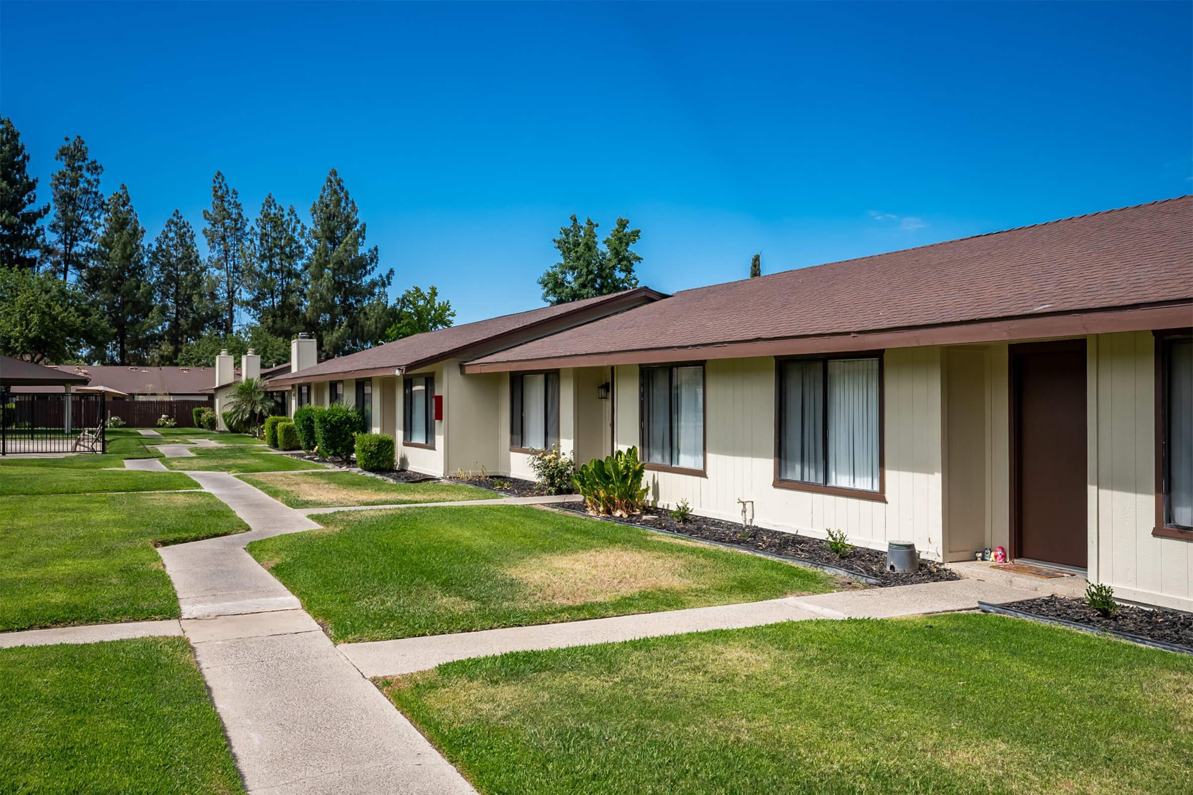 A row of single-story residential buildings with a well-maintained grassy area and pathways. Tall trees are visible in the background under a clear blue sky. The buildings have multiple windows and are surrounded by neatly trimmed shrubs.