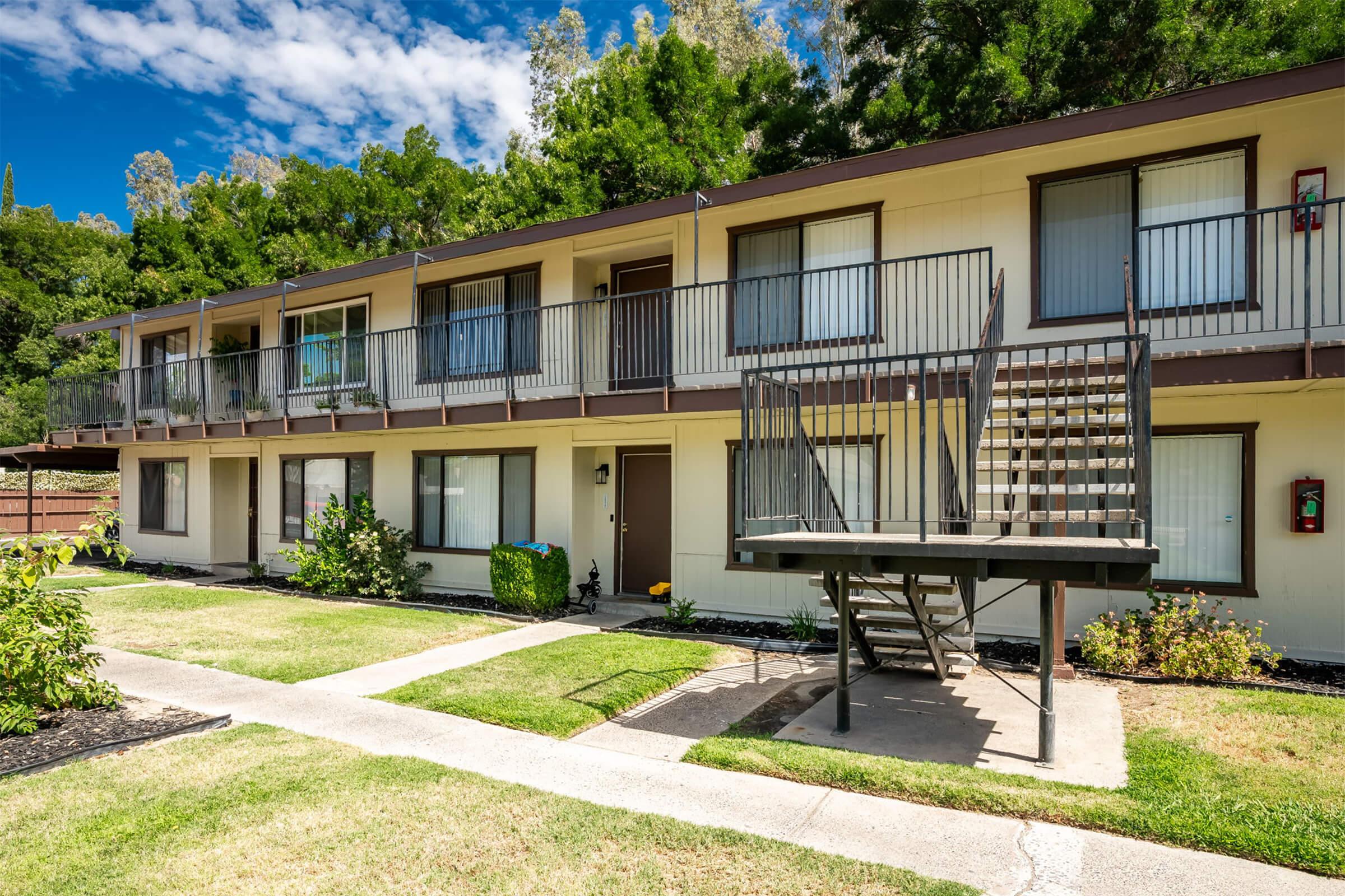 Two-story residential building with multiple balconies and staircases, surrounded by well-maintained grass and shrubs. The exterior features a light-colored facade, large windows, and a concrete pathway leading up to the entrance. Blue sky and trees can be seen in the background.