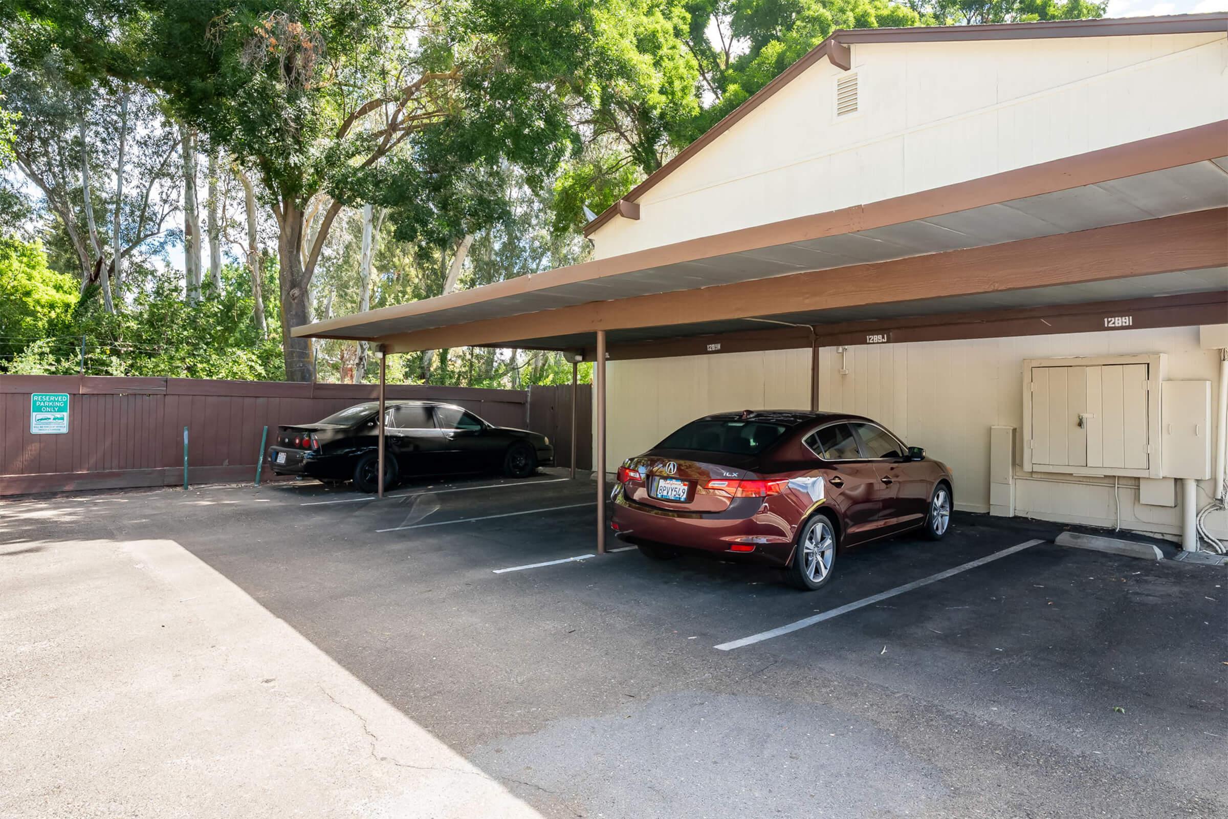 Two parked cars in a shaded outdoor parking area. One car is maroon and the other is black, both under a covered structure. Trees and greenery are visible in the background, enhancing the calm, residential atmosphere. A sign is partially visible on the left side of the image.