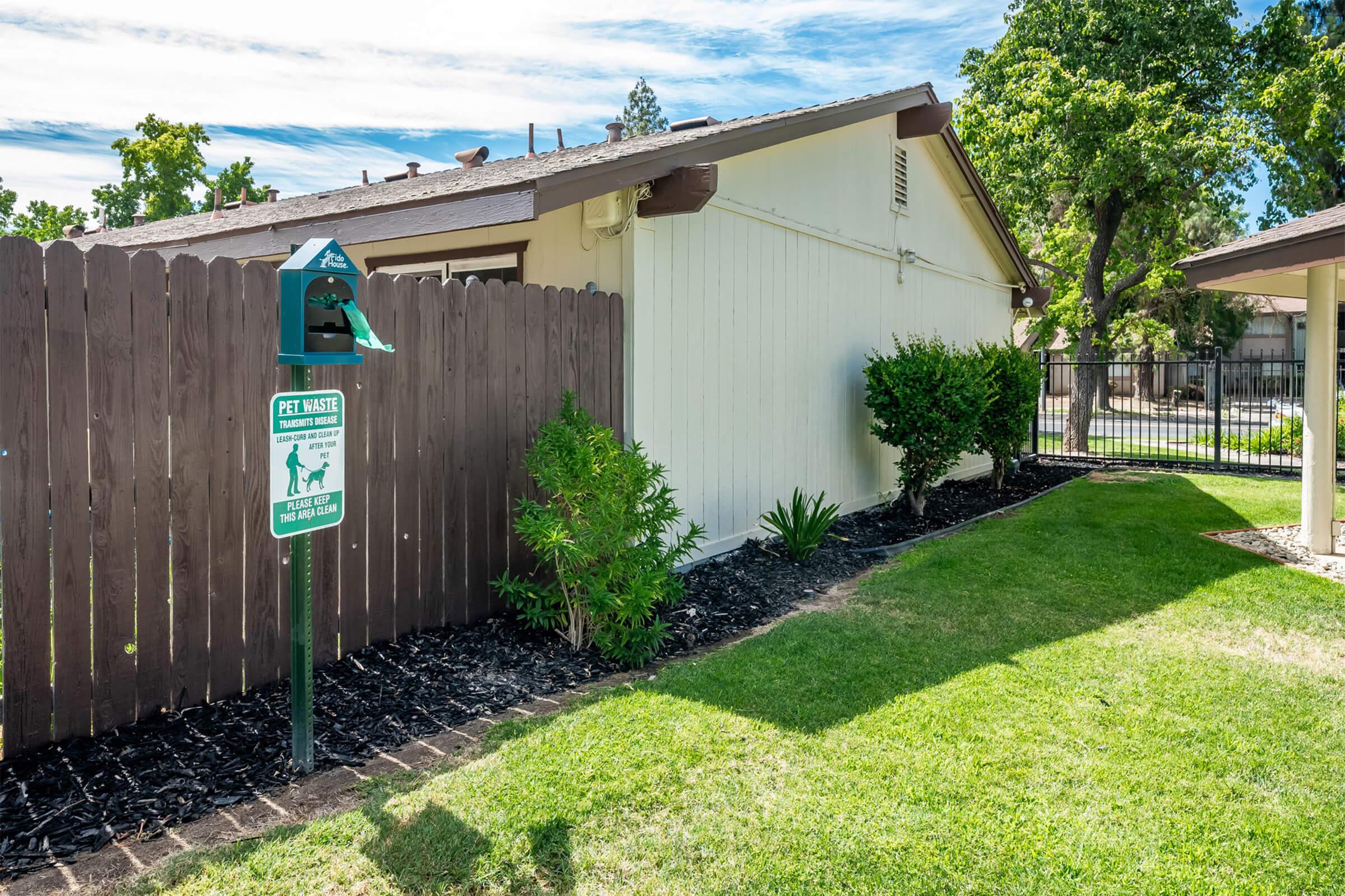 A green pet waste station with a sign is positioned next to a wooden fence. There is a well-maintained grassy area with small shrubs and plants nearby. The house is one story with a light-colored exterior, and a clear blue sky is visible overhead, creating a pleasant outdoor setting.