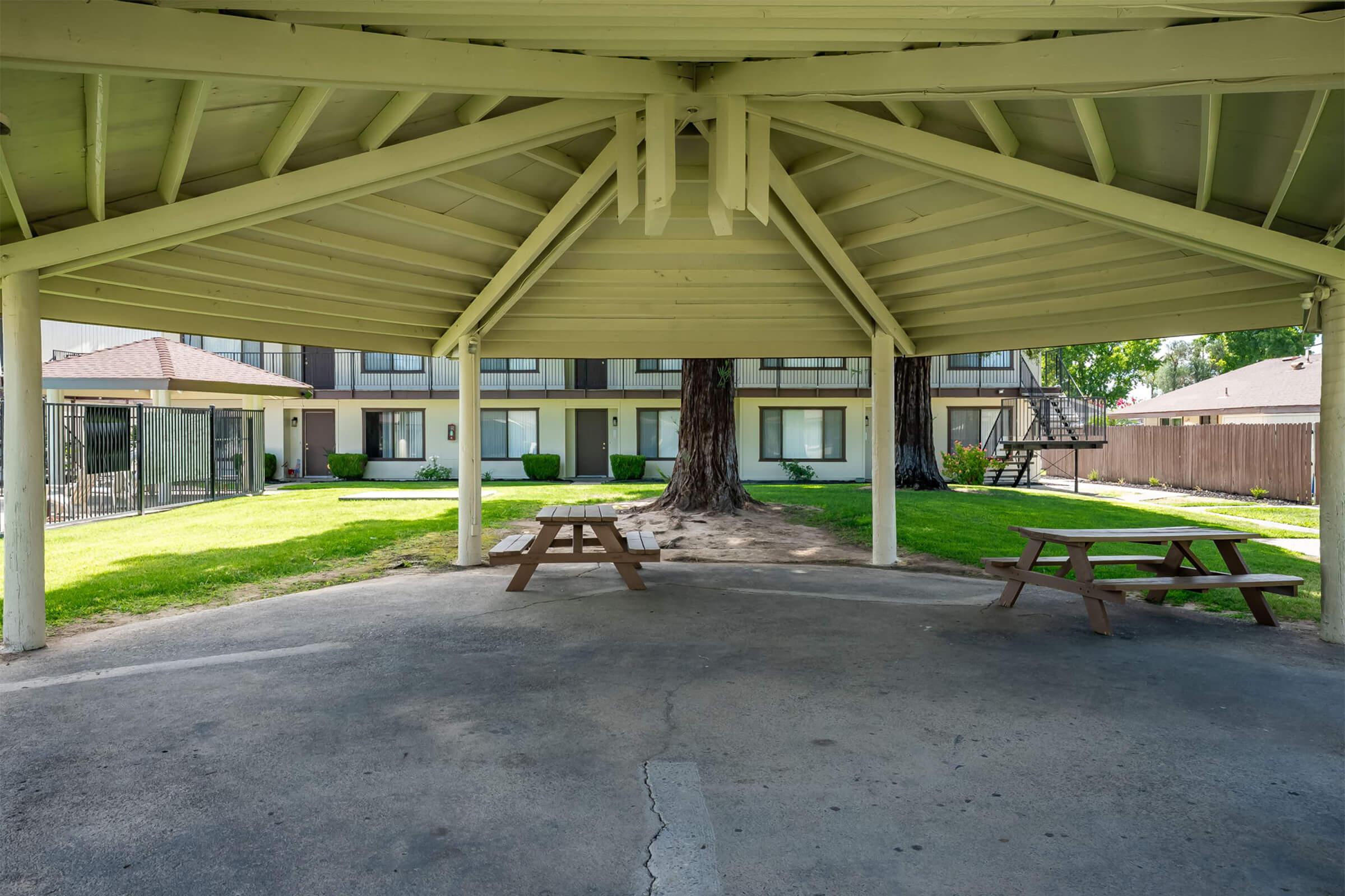 A covered gazebo with a wooden picnic table in a grassy area, surrounded by a courtyard featuring two-story buildings. The structure has a high, slanted roof supported by white pillars, creating a shaded space for relaxation. The area is well-maintained with green lawns and trees.