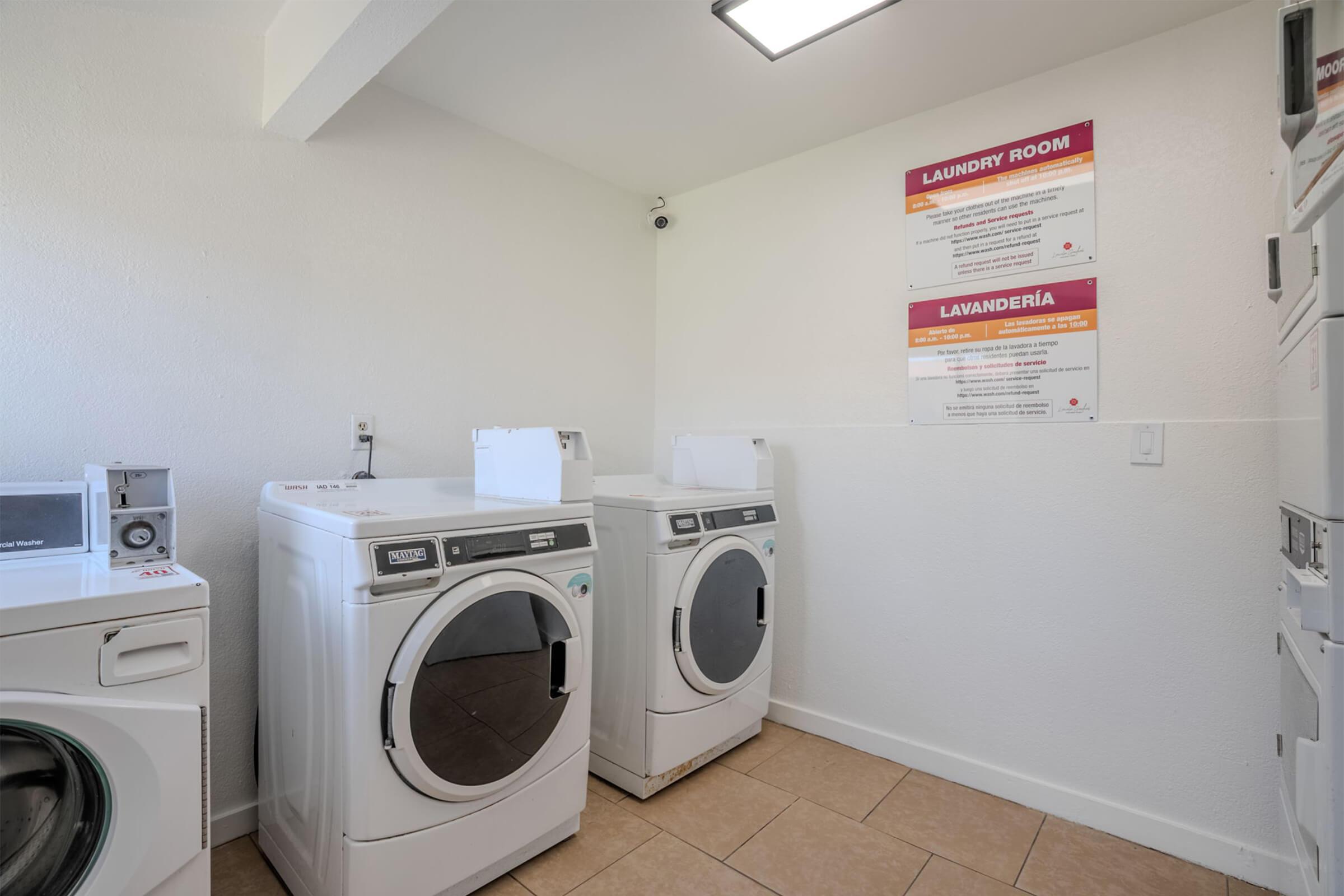 Bright and clean laundry room featuring two front-loading washing machines and stacked dryers. Walls are painted white, and there are two informational signs in English and Spanish about laundry room guidelines. The floor is tiled, and the overall space is well-lit.
