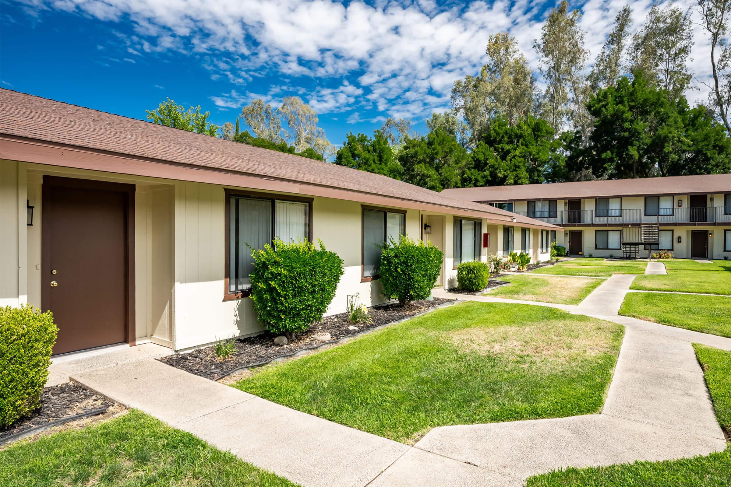 A well-maintained outdoor view of a low-rise apartment complex, featuring multiple units with brown doors, neatly trimmed bushes, and a well-kept lawn. The scene is bright with a blue sky and scattered clouds, showcasing a clean, inviting atmosphere with concrete pathways between the units.