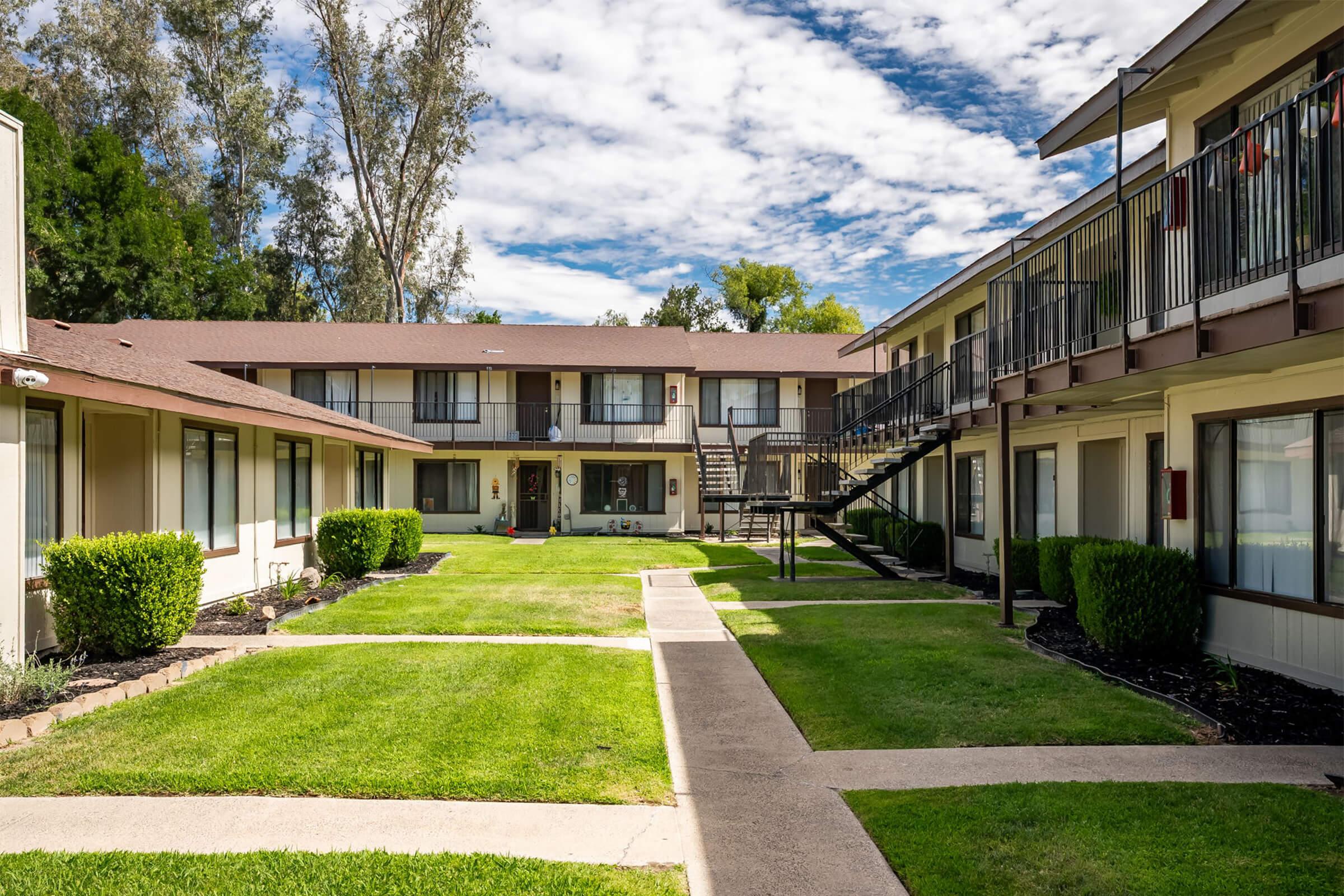 A view of a landscaped courtyard featuring two-story apartment buildings. The area is neatly maintained with green grass and hedges, surrounded by walkways. Stairs lead to the upper level of one building, under a partly cloudy blue sky.