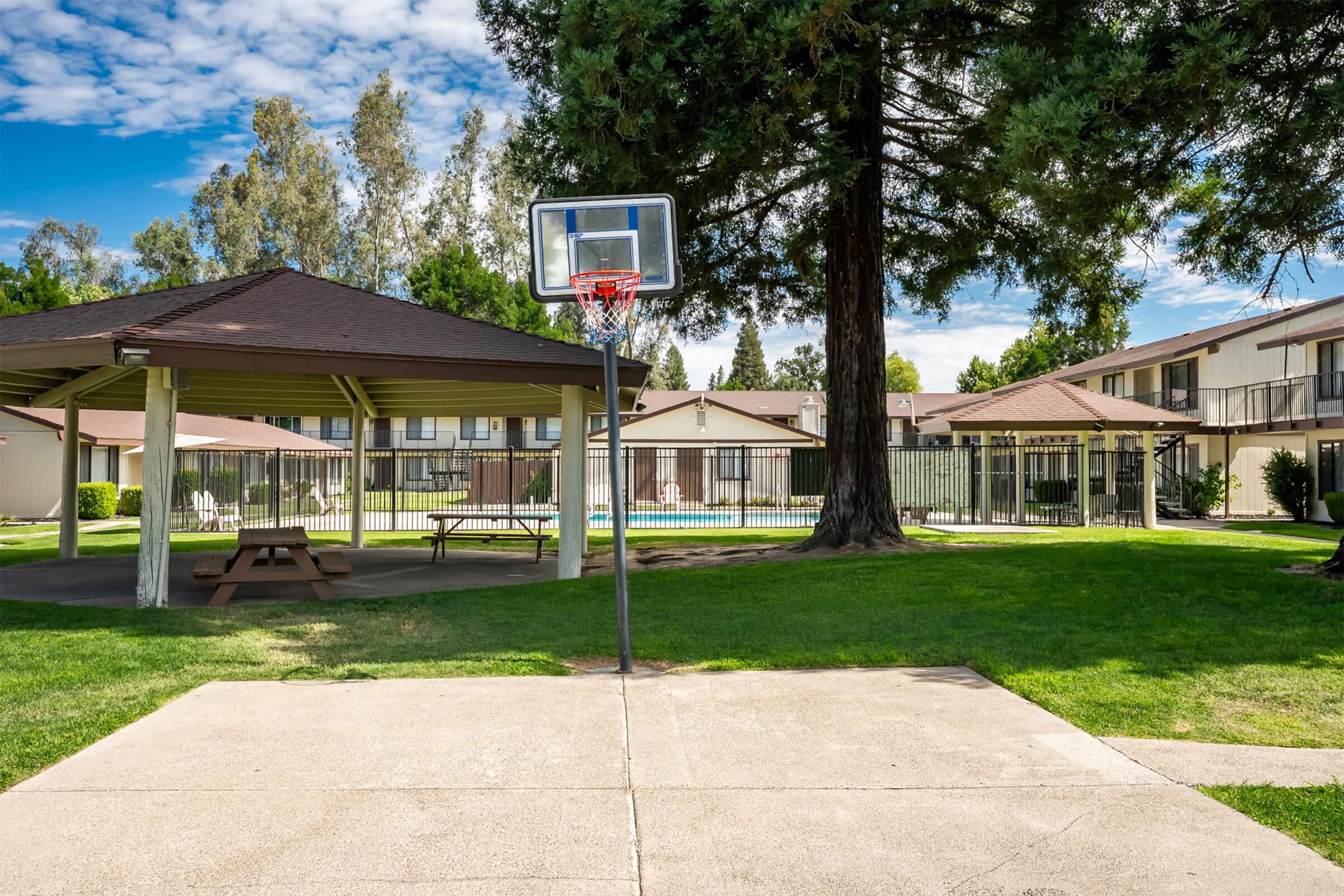 A view of a basketball hoop next to a shaded gazebo and a grassy area. Behind the hoop, there are several residential buildings and a fenced-in swimming pool. The sky is blue with some clouds, and there are tall trees in the background, creating a pleasant outdoor environment.