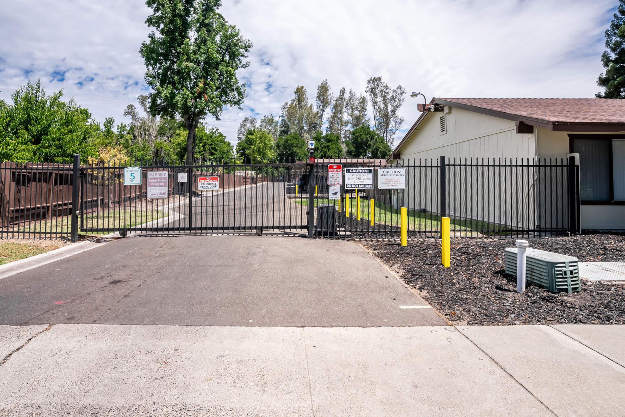 A closed security gate at an entrance with various warning signs. Lush green trees and shrubs line the pathway, which is paved and well-maintained. A building can be seen to the right, and there are yellow bollards on either side of the gate. The sky is partly cloudy.