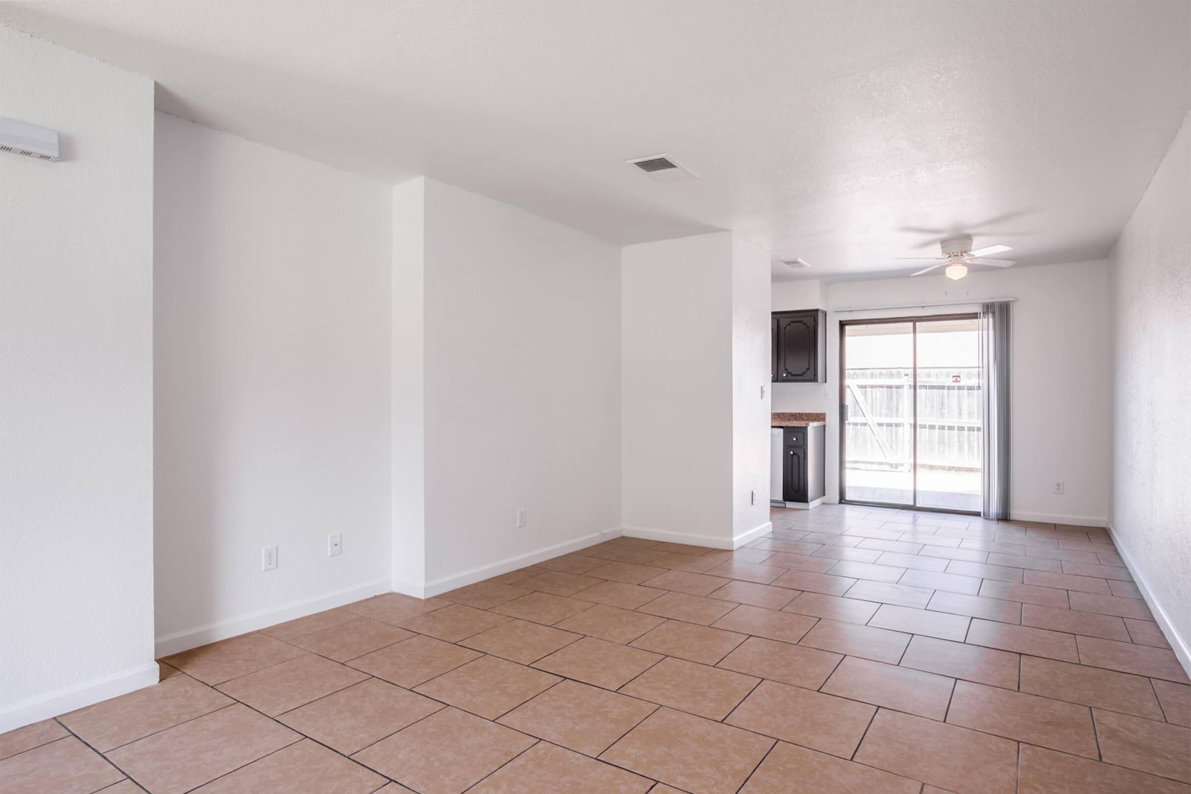 A spacious, empty living room featuring light-colored walls and large tile flooring. One side has a sliding glass door leading to an outdoor area, while the other side shows a small kitchen nook with dark cabinetry. Natural light fills the room, creating a bright and airy atmosphere.