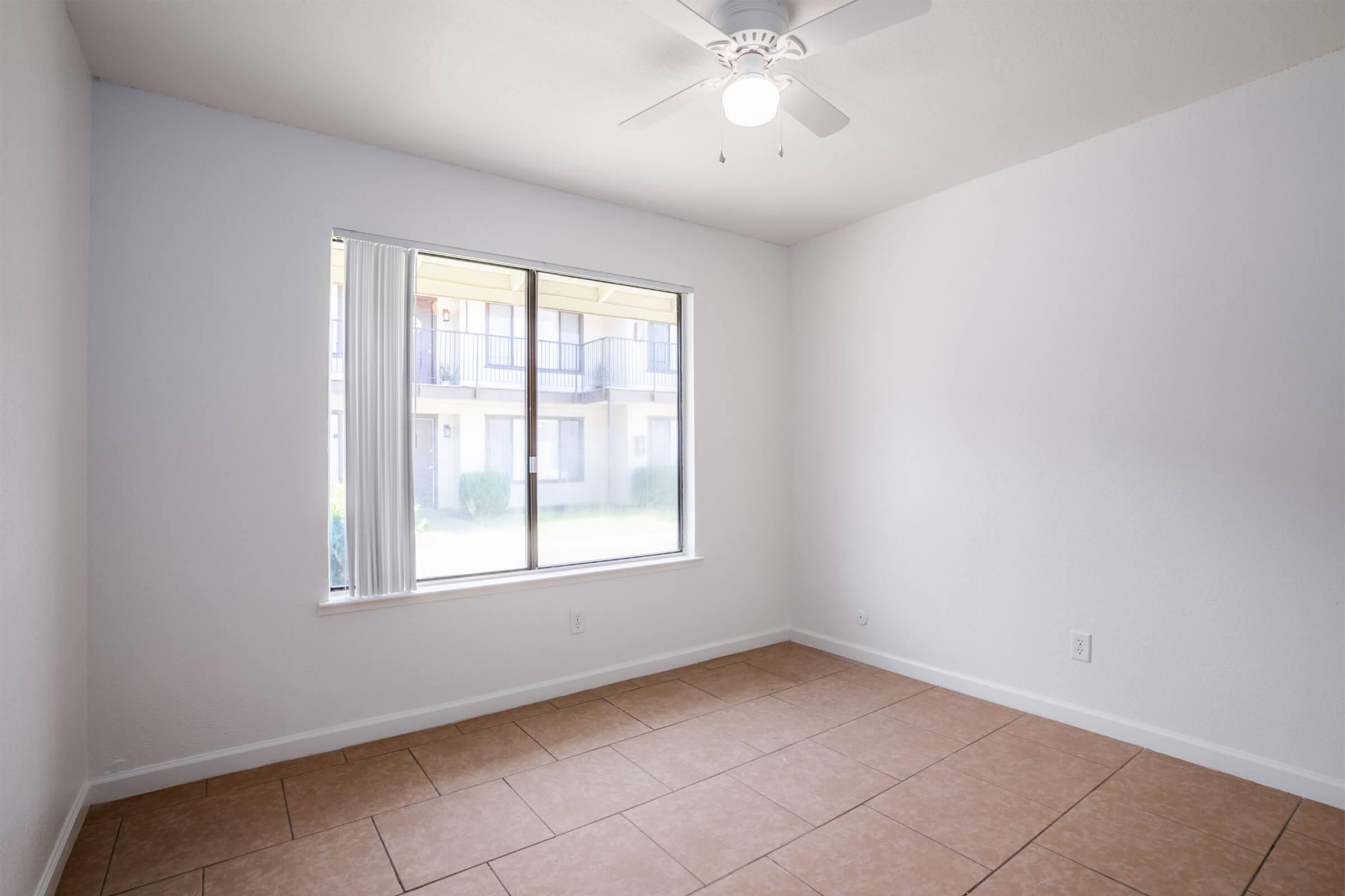 A bright, empty room with white walls and a ceiling fan. The large window offers a view of an outdoor area with balconies. The floor is tiled with neutral-colored tiles, creating a clean and inviting space.