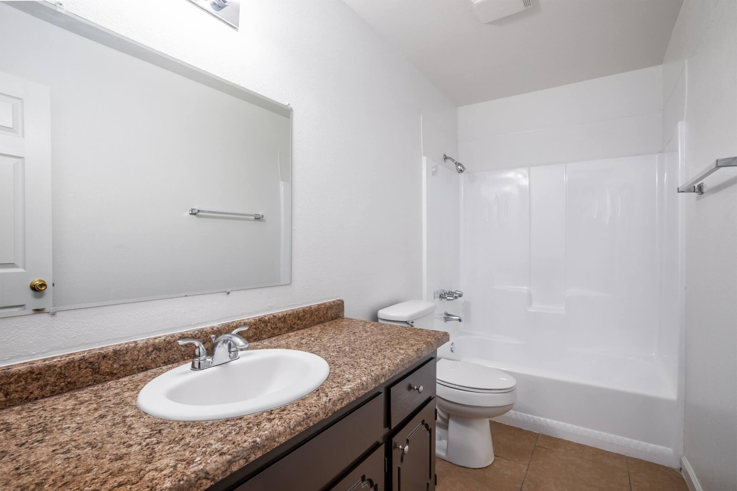 A clean and modern bathroom featuring a granite countertop sink, a large mirror above the sink, a white bathtub and shower combination, and a toilet. The walls are painted white, and the floor is tiled in a neutral tone.