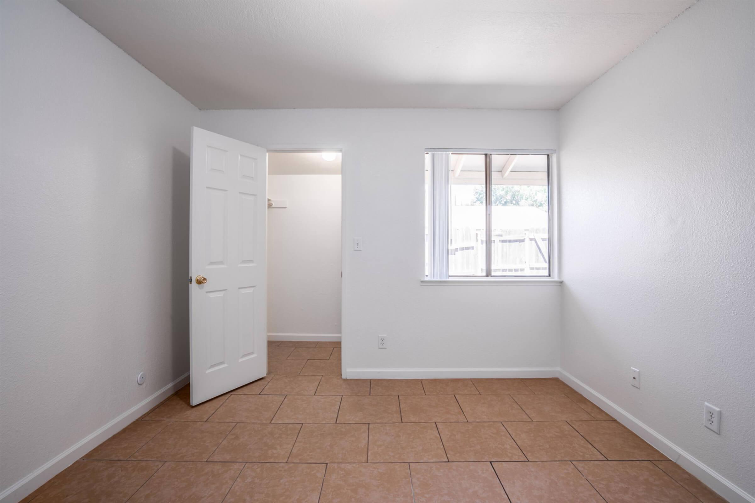 An empty room with white walls and a tan tile floor. A closed door on the left leads to another space, while a window on the right allows natural light to enter. The room is well-lit and uncluttered, creating a simple and clean appearance.