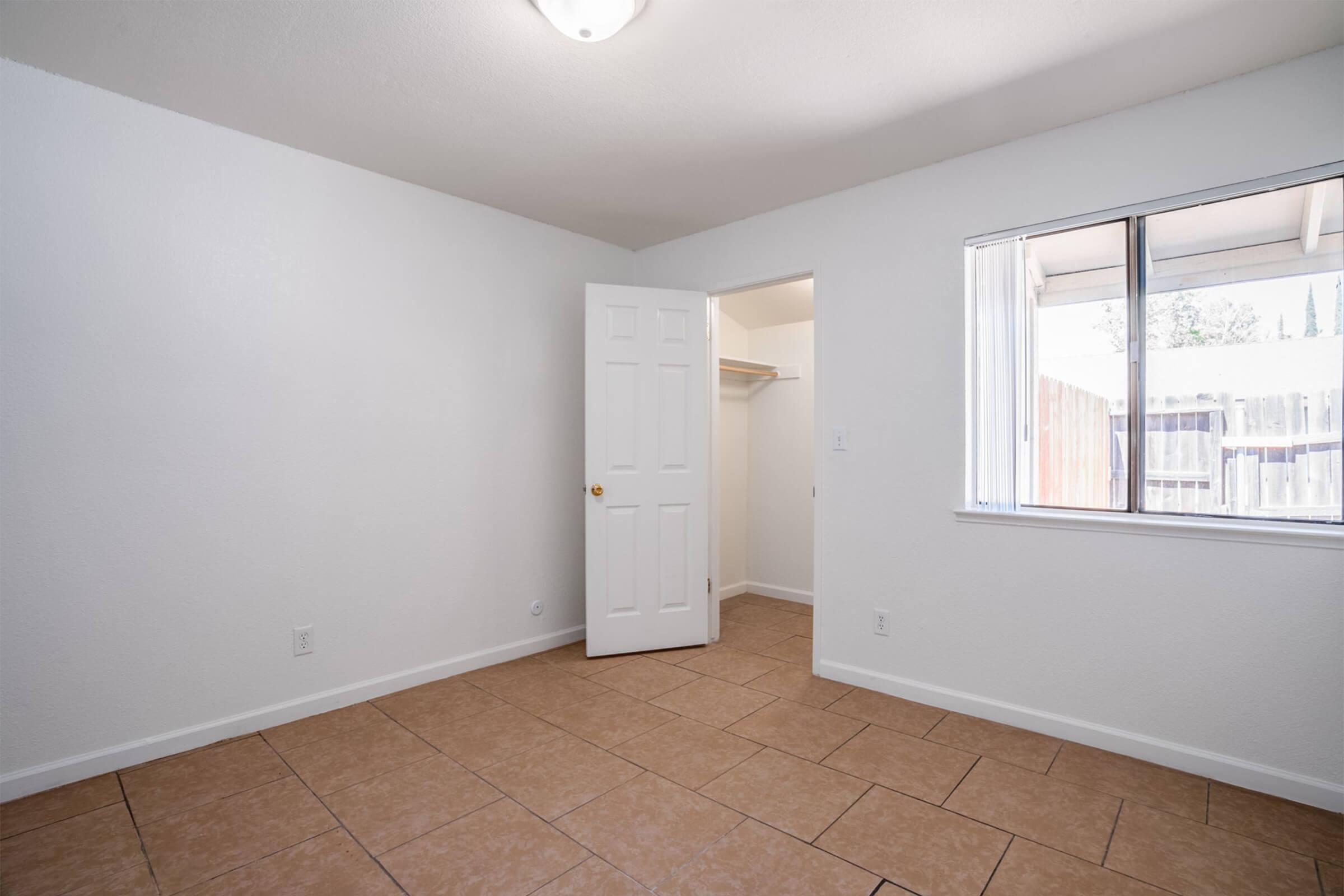 A spacious, empty room with white walls and a light-colored tiled floor. There is a closed door leading to a closet, and a window allowing natural light to come in. The overall atmosphere is bright and minimalistic, suitable for a variety of purposes such as a bedroom or office.