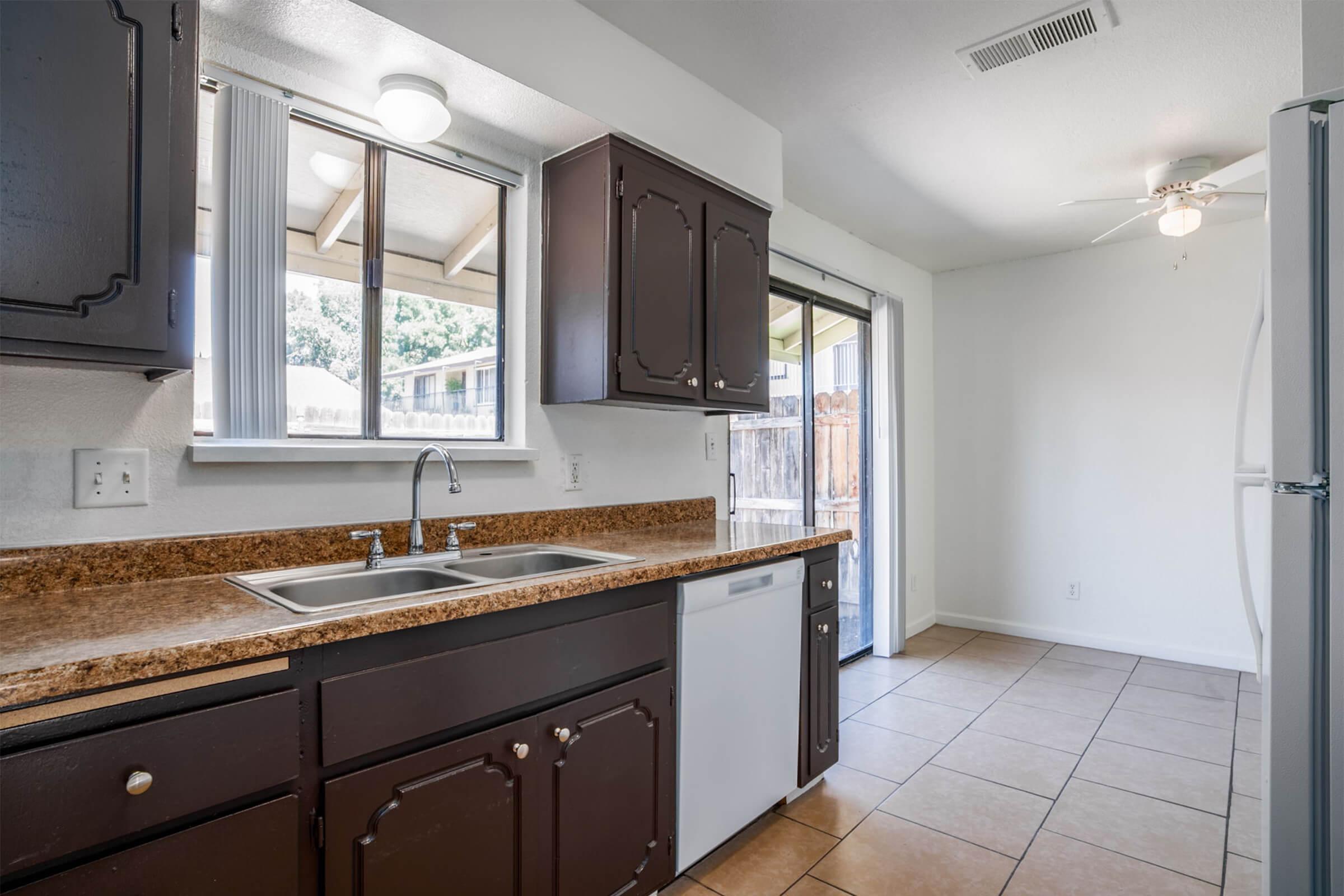 Spacious kitchen with dark wood cabinetry, a double sink, and modern appliances. Natural light pours in through a window and sliding door, leading to an outdoor area. The countertops are a light speckled design, and the flooring is tiled in a neutral color, creating a warm and inviting atmosphere.