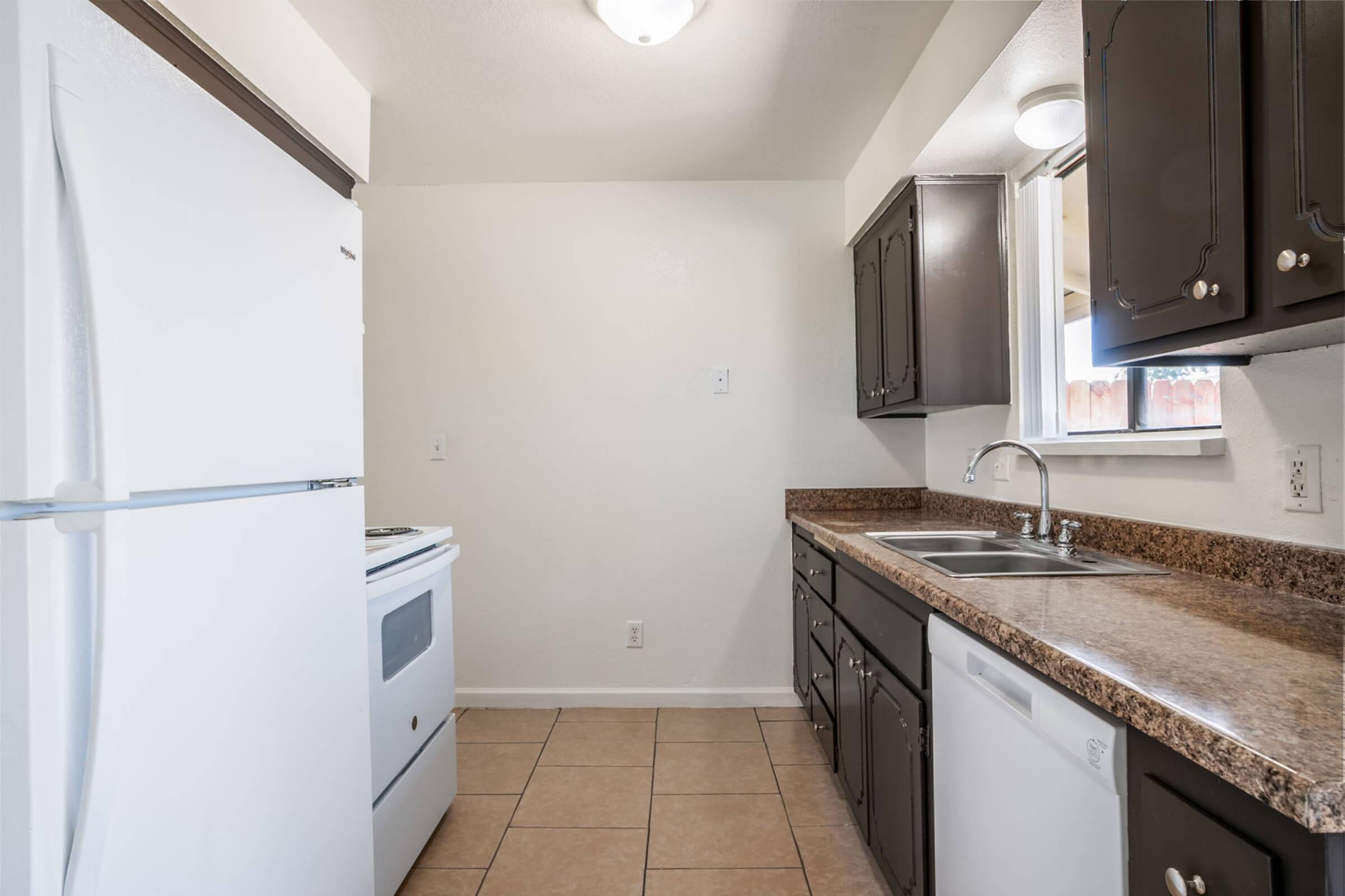 A clean and simple kitchen featuring white appliances, including a refrigerator and stove. The countertops are a light brown speckled design, and there are dark cabinets above and below the sink. The walls are painted a light color, creating a bright and spacious feel. Tile flooring completes the space.