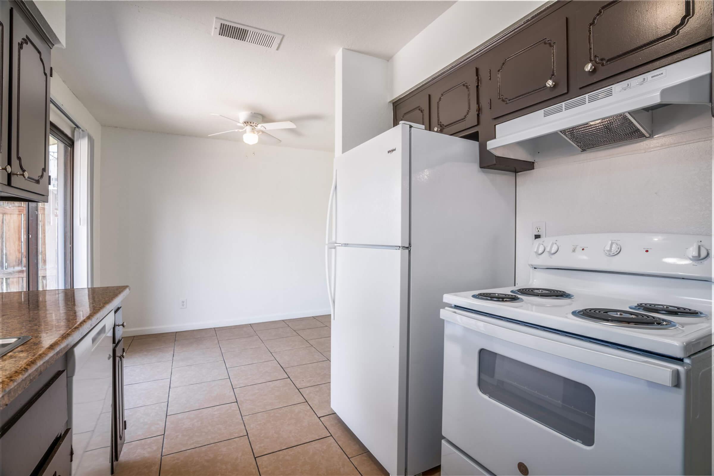 A clean, modern kitchen featuring a white refrigerator, a white stove with an oven, and dark wood cabinets. The space has tile flooring and a ceiling fan, with a bright, well-lit atmosphere enhanced by a window.