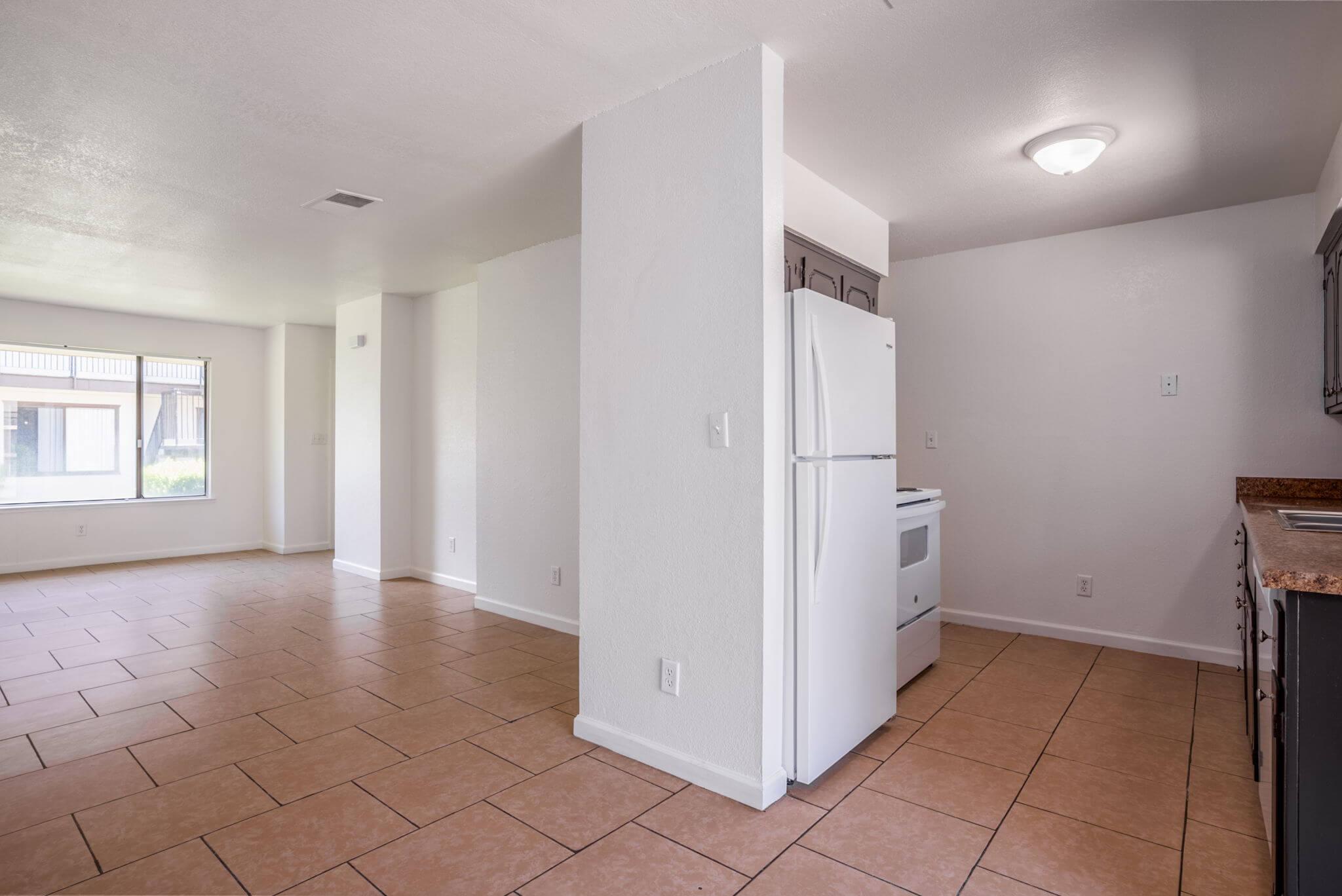 Empty room with tiled flooring, featuring a kitchenette with a white fridge and stove. There are windows allowing natural light, and the walls are painted in a light color, creating a spacious atmosphere. The layout suggests a combined living and dining area.