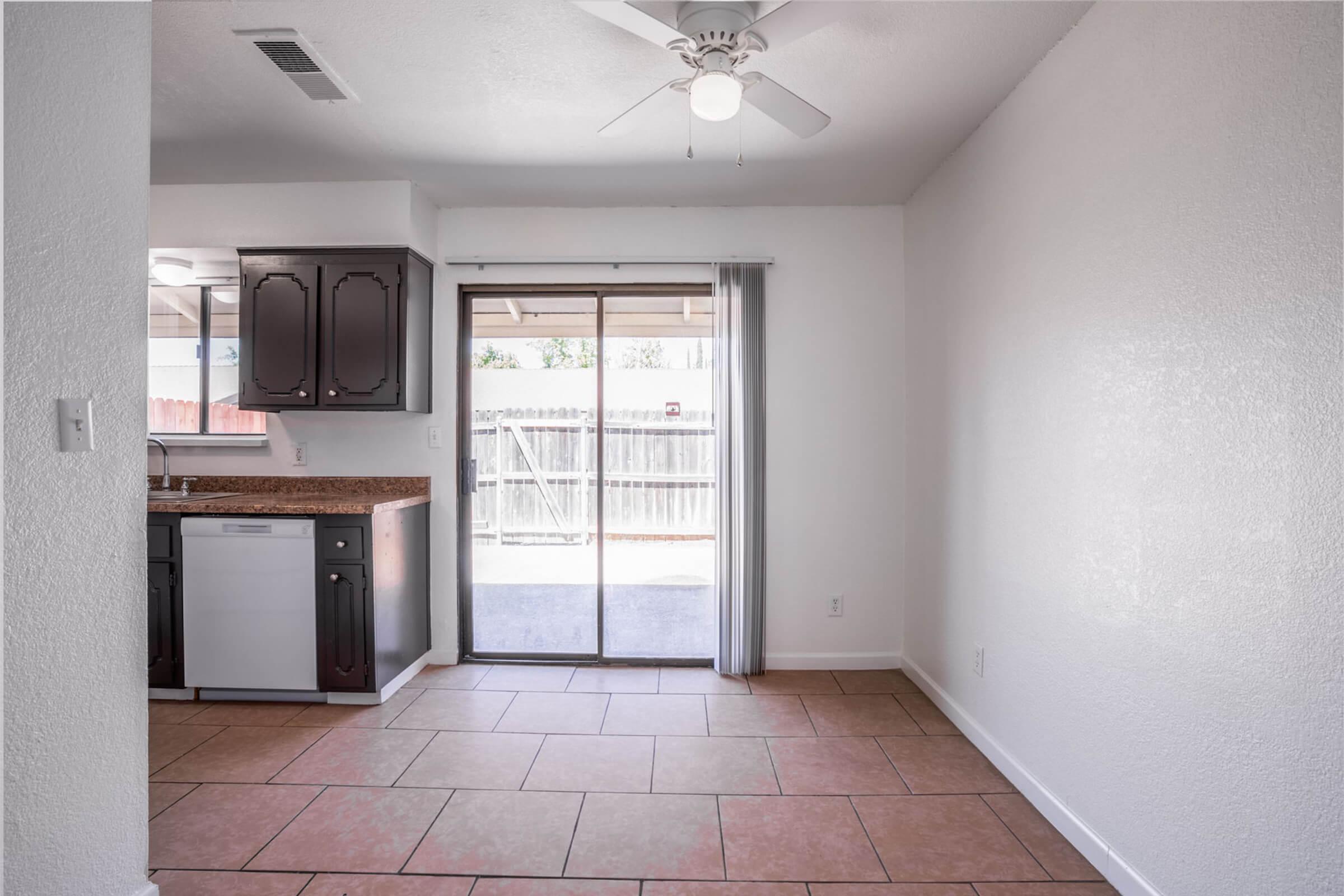 A bright and clean kitchen and living area with tiled flooring, featuring a ceiling fan, sliding glass doors leading to an outdoor space, and dark wood cabinets. The kitchen includes a dishwasher and a countertop, creating an open and inviting atmosphere.