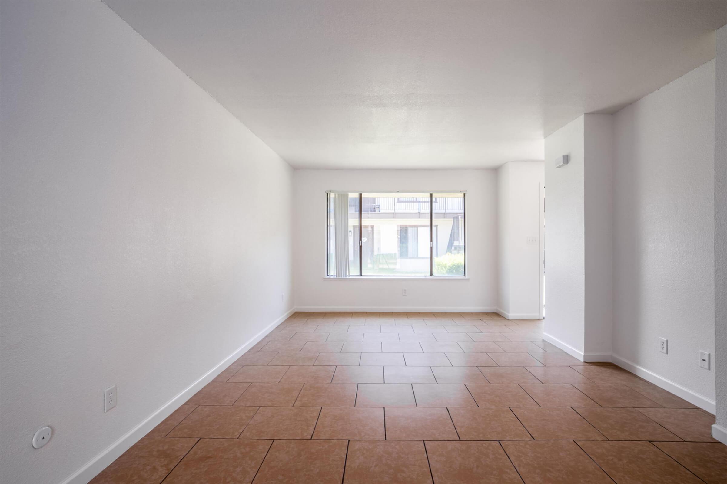 A spacious, empty room with light-colored walls and a tiled floor. There is a large window allowing natural light to enter, providing a view of an outdoor space or courtyard. The room has a clean and minimalist appearance, suitable for various layouts.