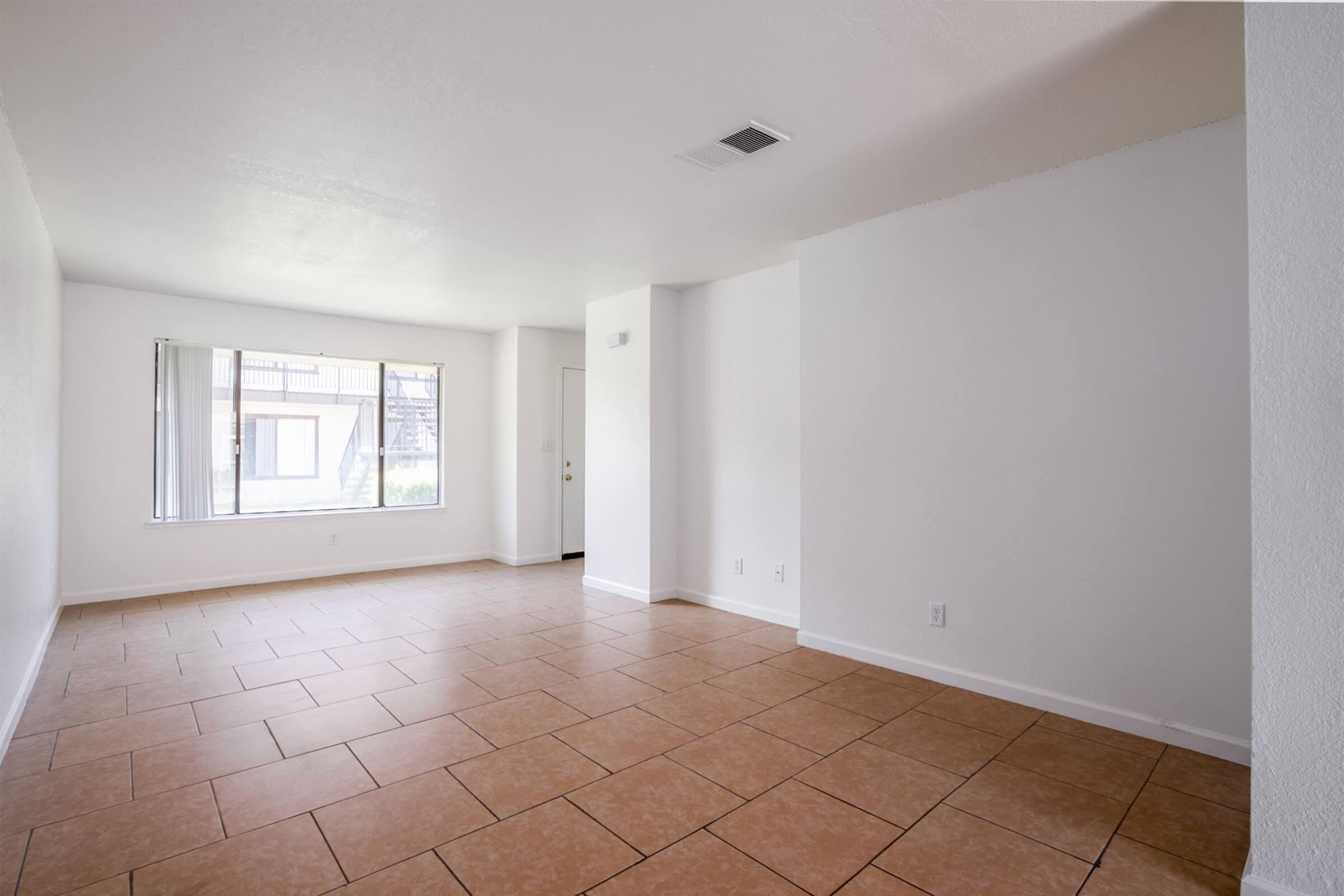 Empty room with white walls and large windows allowing natural light. The floor is covered with light brown tiles. There are no furnishings or decorations, creating a spacious and minimalist atmosphere. A door is visible in the corner, leading outside.
