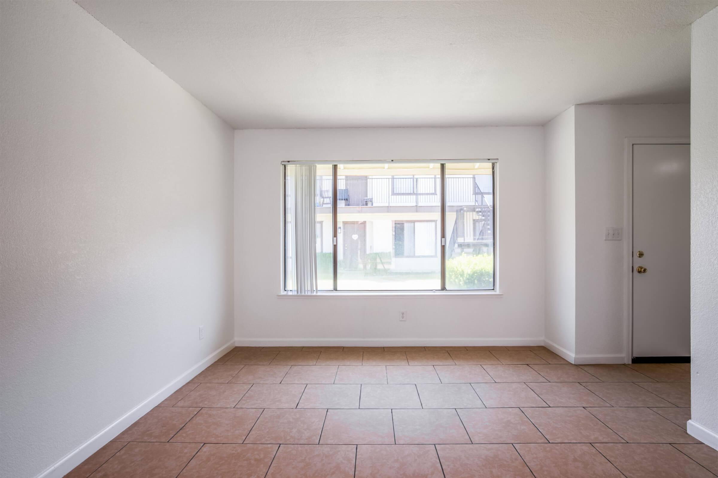 An empty room with white walls and tiled flooring, featuring a large window that provides natural light. The window looks out onto a grassy area and an apartment complex with a balcony. There is a door on the right side of the image.