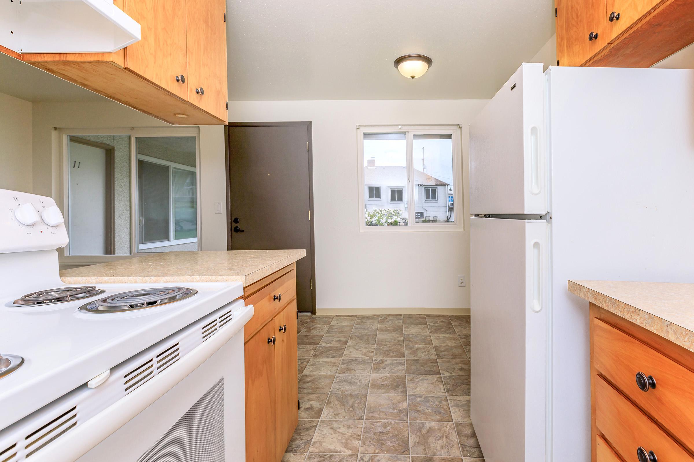 A bright kitchen interior featuring wooden cabinetry, a white stove, a white refrigerator, and tiled flooring. The kitchen has a window with a view to the outside, allowing natural light to enter, and a door leading to another room. The space is clean and modern, providing an inviting atmosphere.
