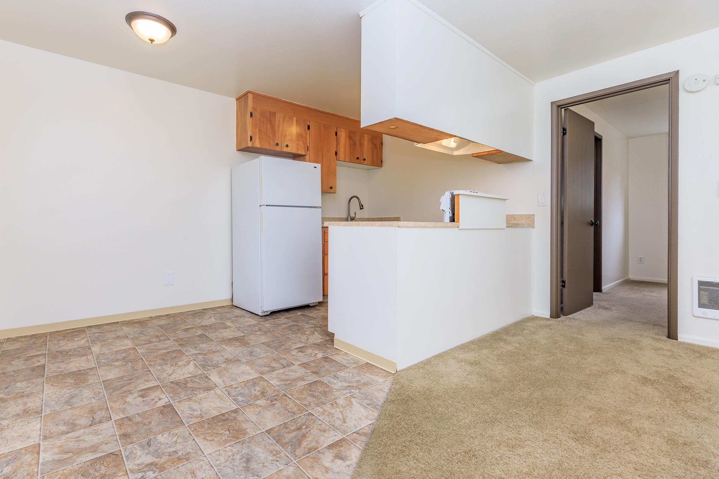 A small, modern kitchen featuring a white refrigerator, wooden cabinets, and a sink. The flooring is a patterned tile design, leading into a carpeted living area. A doorway on the right leads to another room, and there is overhead lighting in the kitchen space. The walls are painted a light color, creating a bright atmosphere.